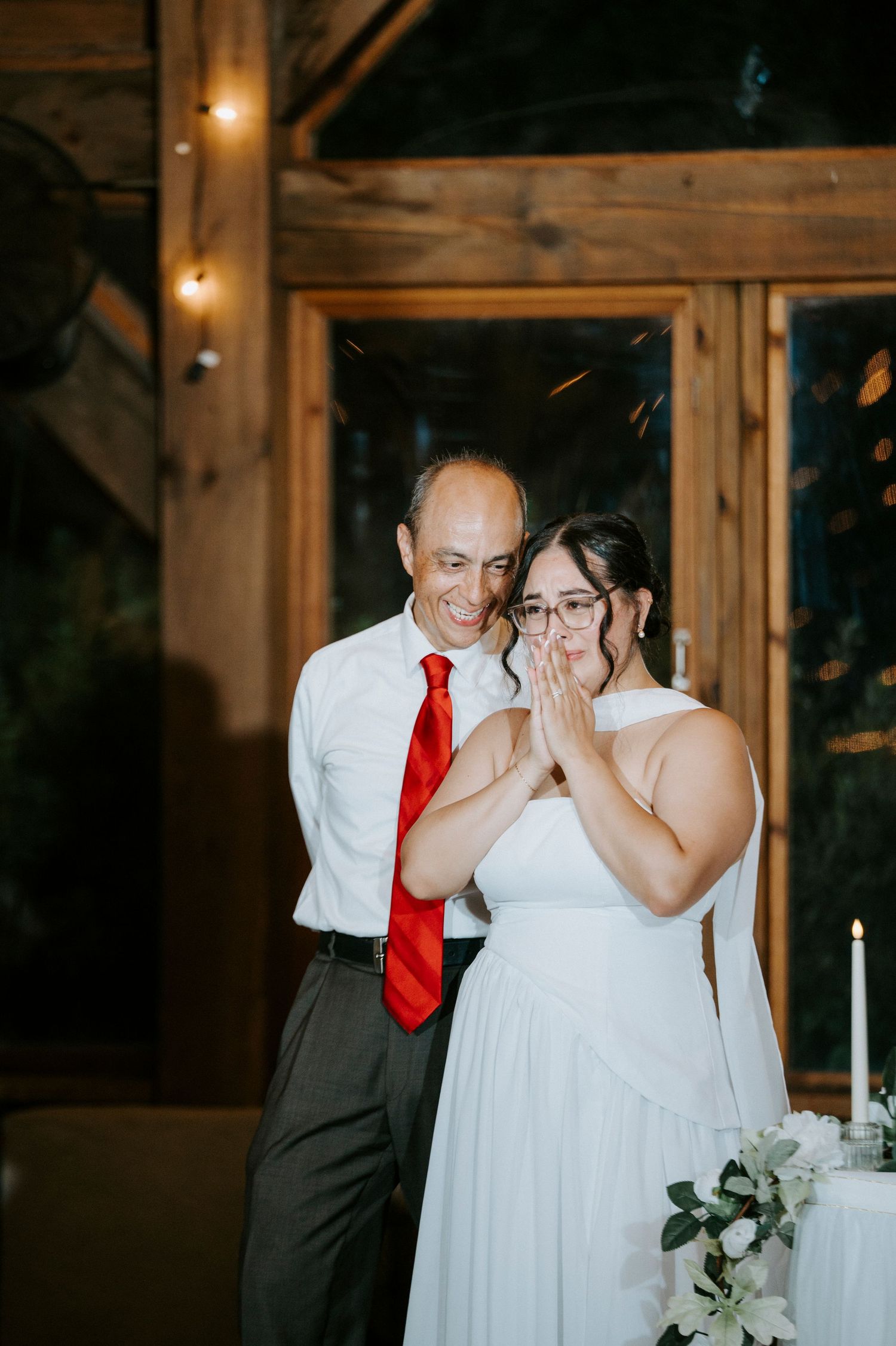 A touching wedding reception moment shows a couple sharing an emotional embrace in a rustic wooden venue with warm lighting.