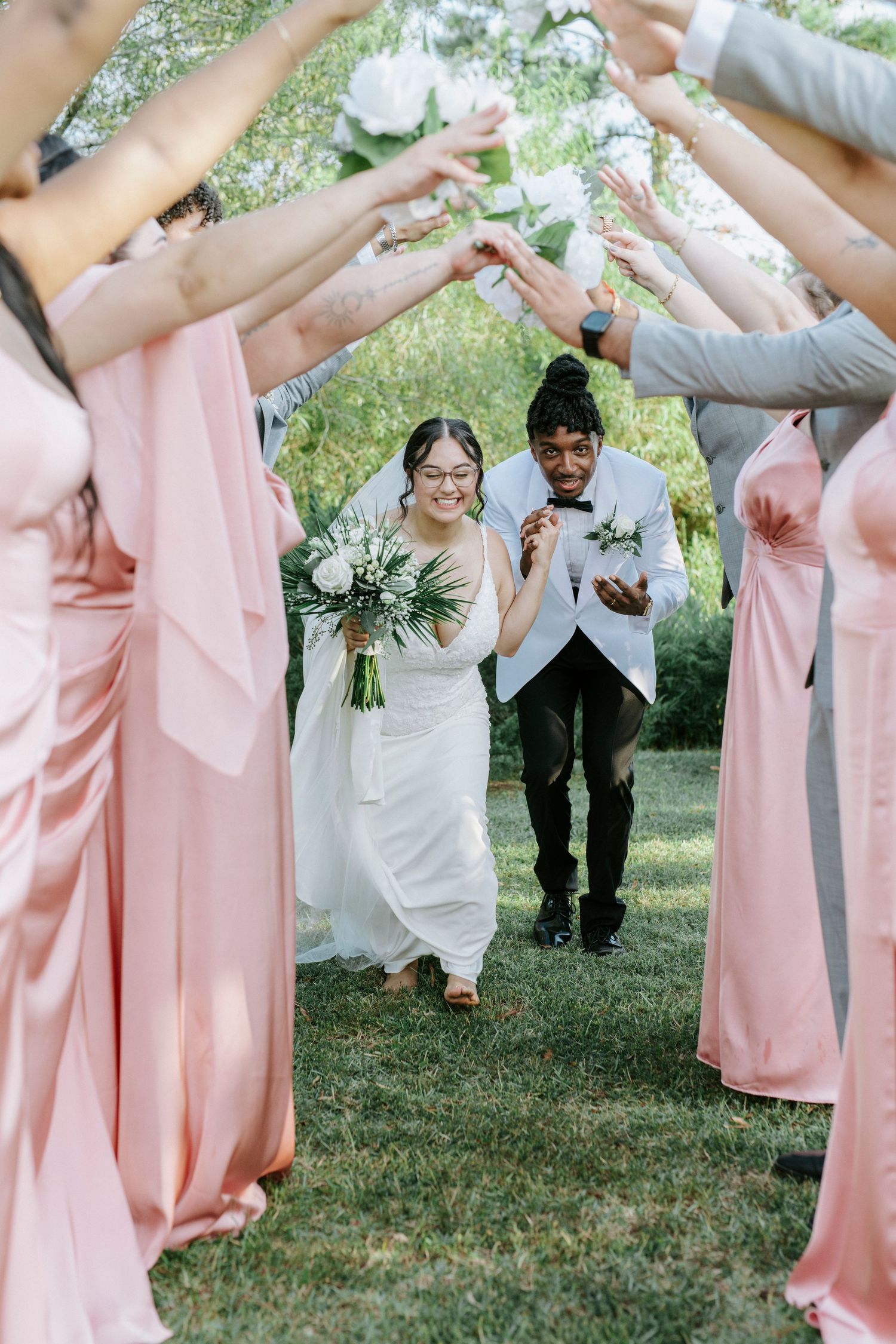 Wedding party members in pink bridesmaid dresses and suits form a tunnel on grass for newlyweds to walk through.