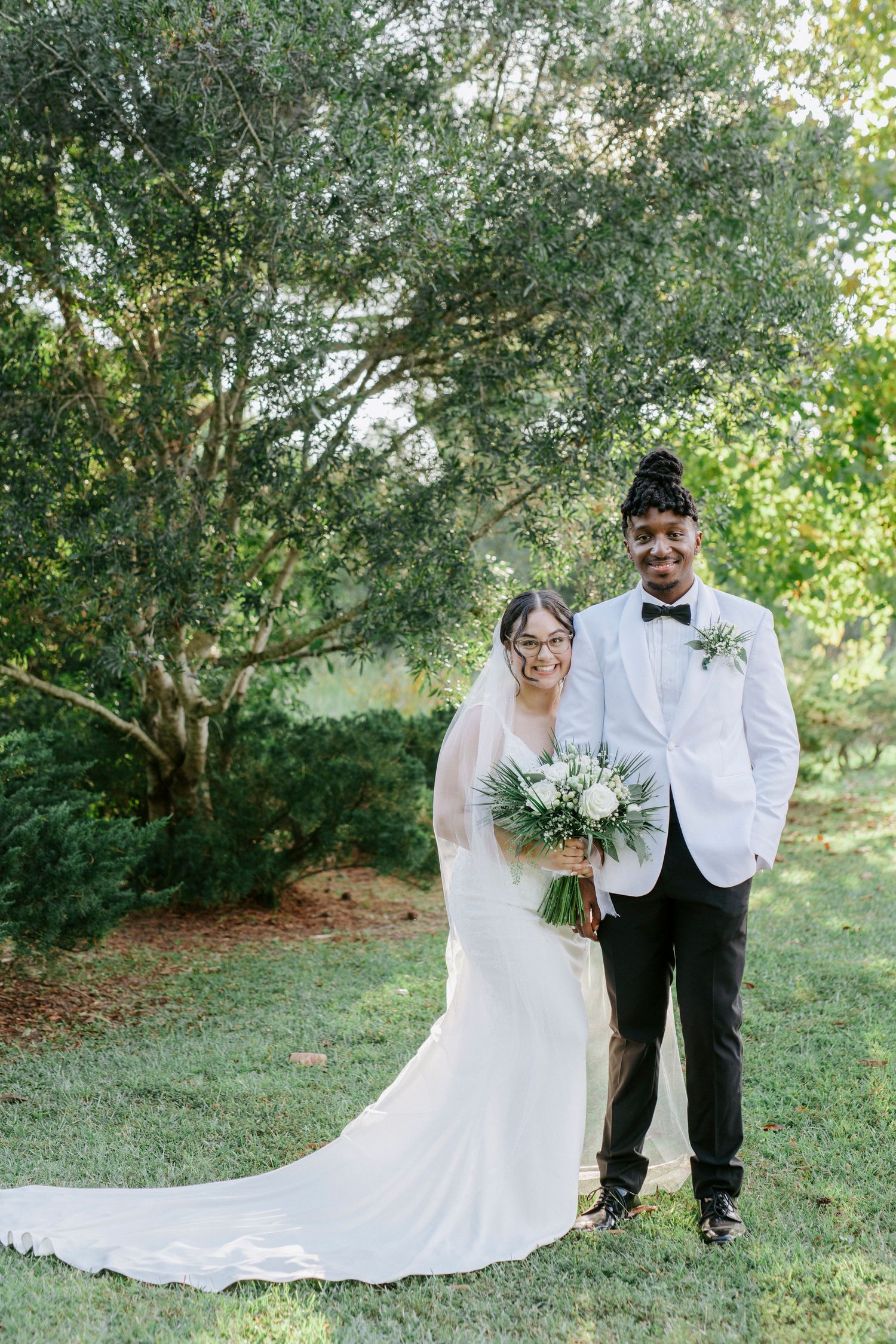 A couple in wedding attire poses outdoors with a white bridal gown and bouquet against a lush garden backdrop.