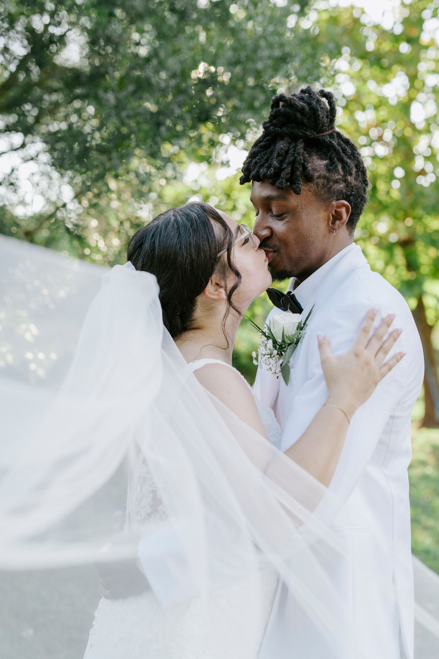 A romantic wedding couple shares tender kisses in a garden setting with flowing white veil and natural greenery.