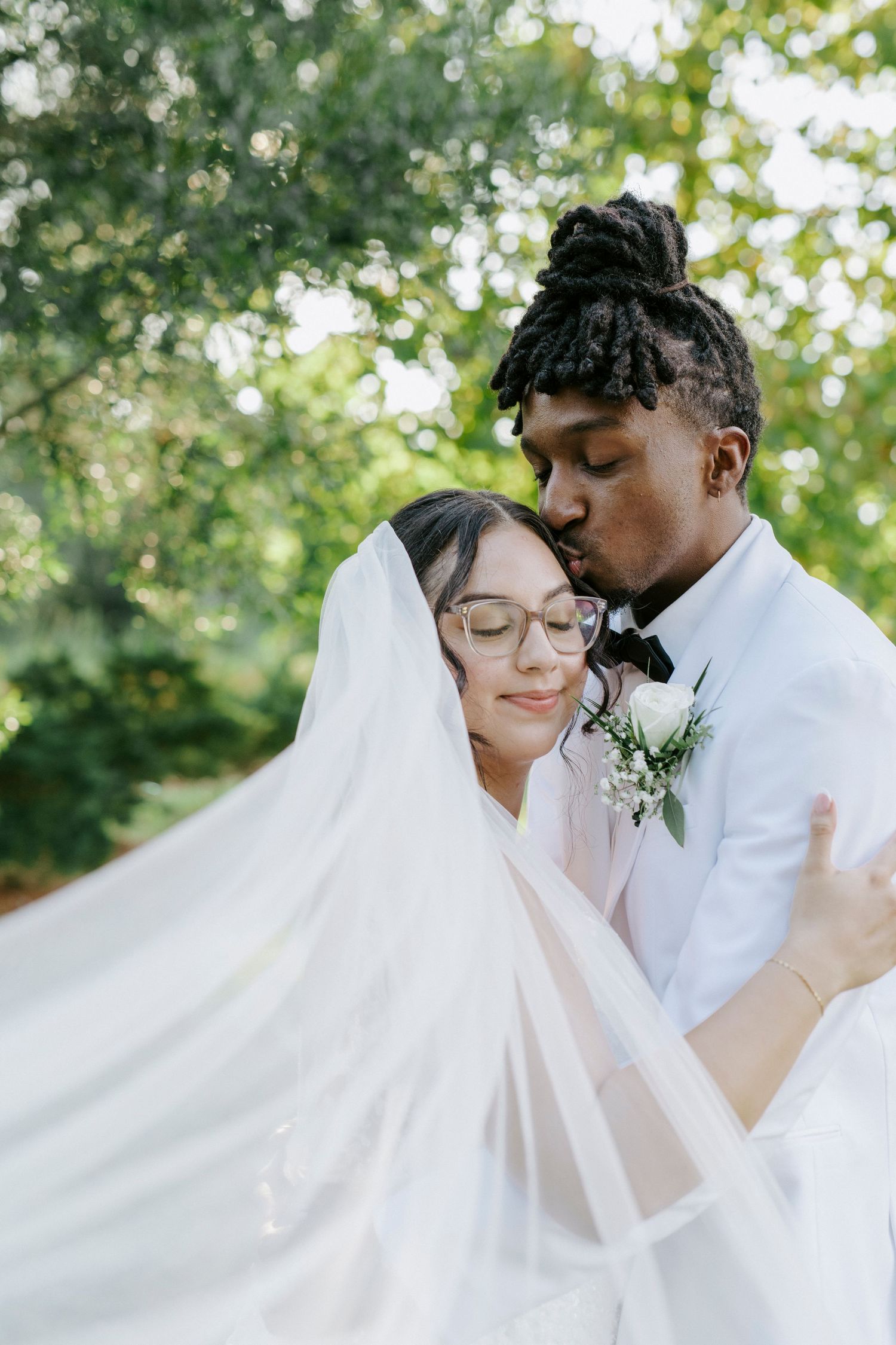 Romantic wedding couple shares intimate moments outdoors with flowing white veil and floral details in natural lighting.