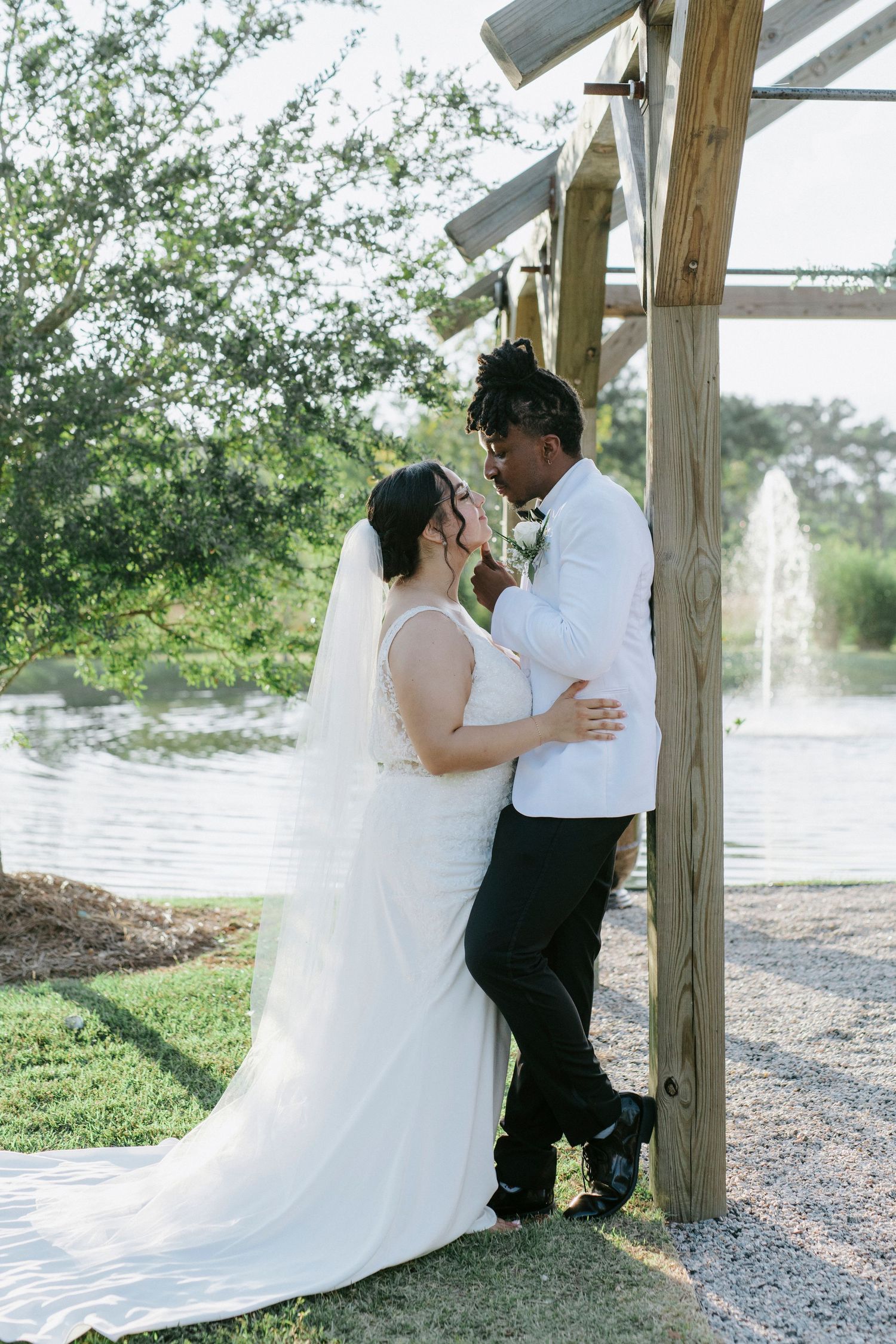 A romantic wedding couple shares intimate moments under a wooden gazebo by a serene waterfront setting.