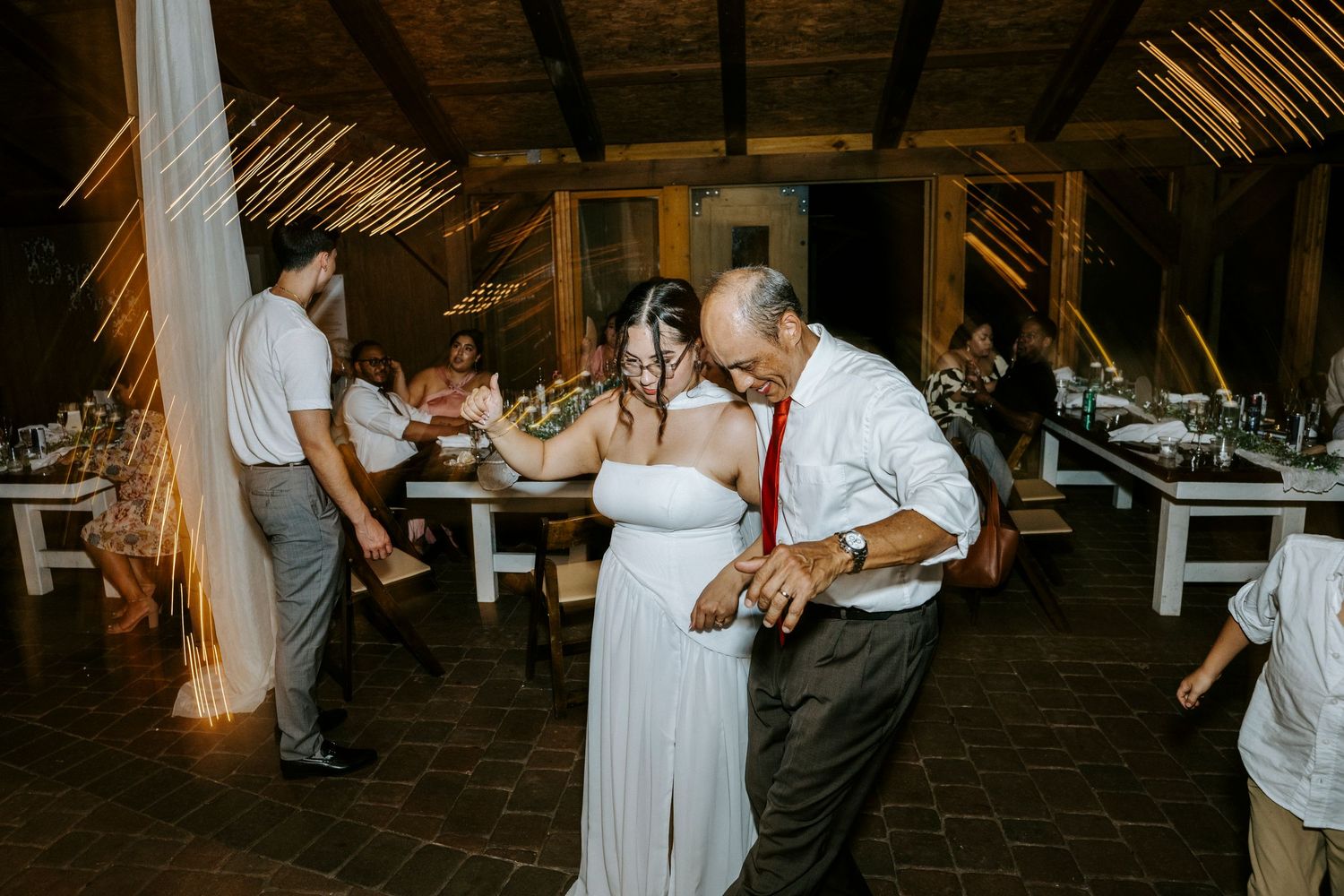 Couples dance together during an evening celebration in a rustic wooden barn venue with warm lighting.