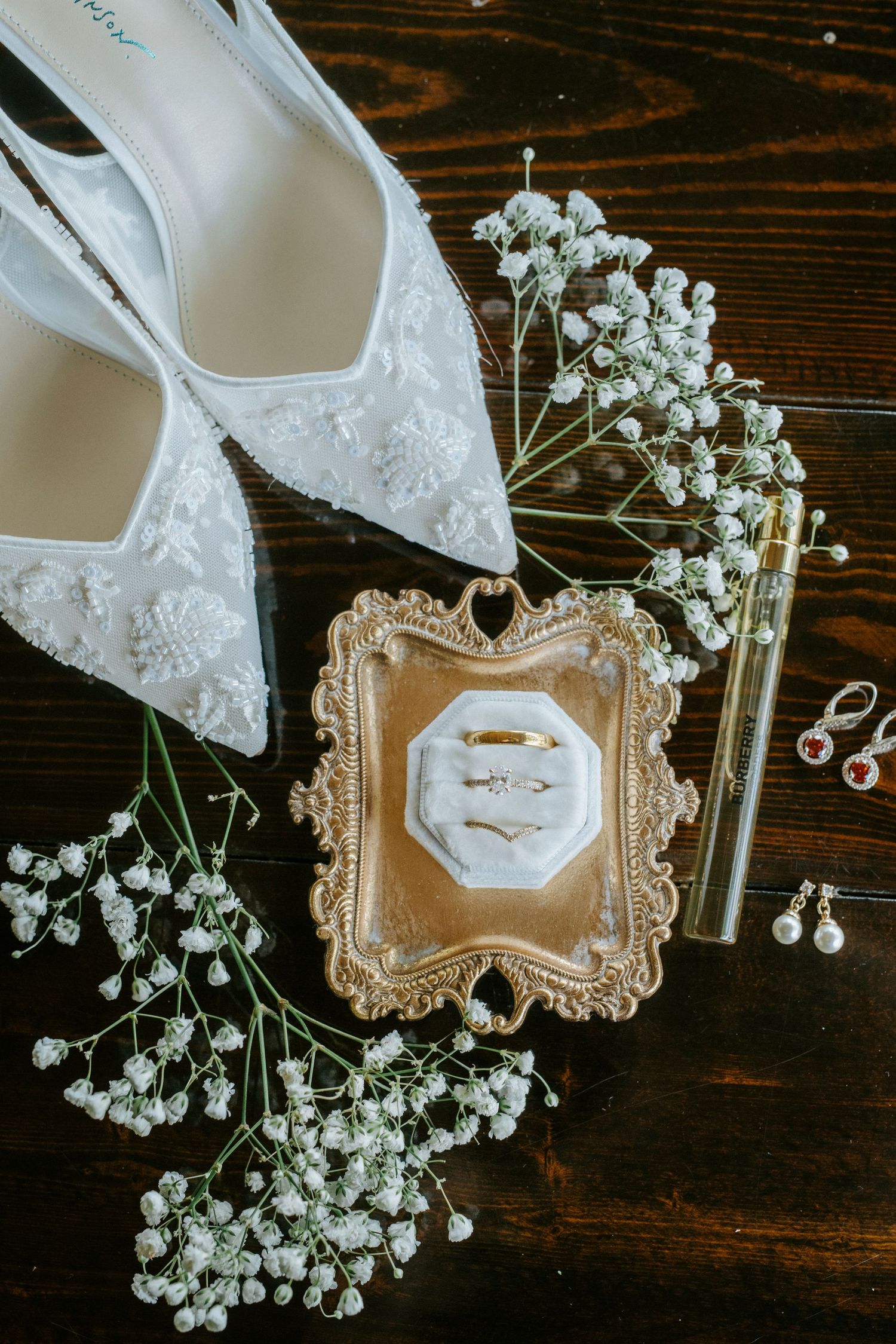 Elegant bridal accessories including white lace shoes, baby's breath flowers, and vintage-style frames on dark wooden surface.