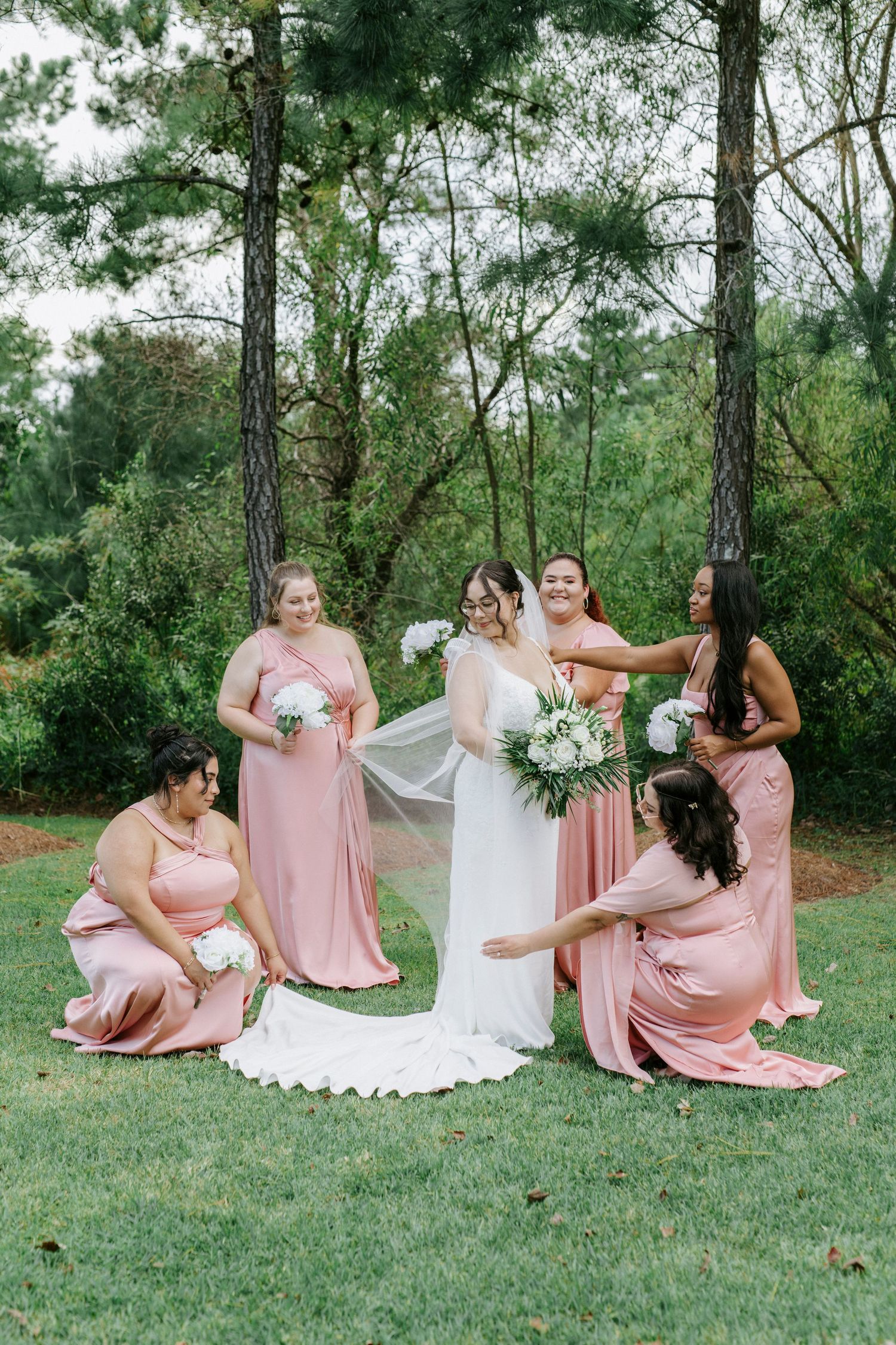A bridal party in pink dresses poses together on a grassy lawn surrounded by tall pine trees.