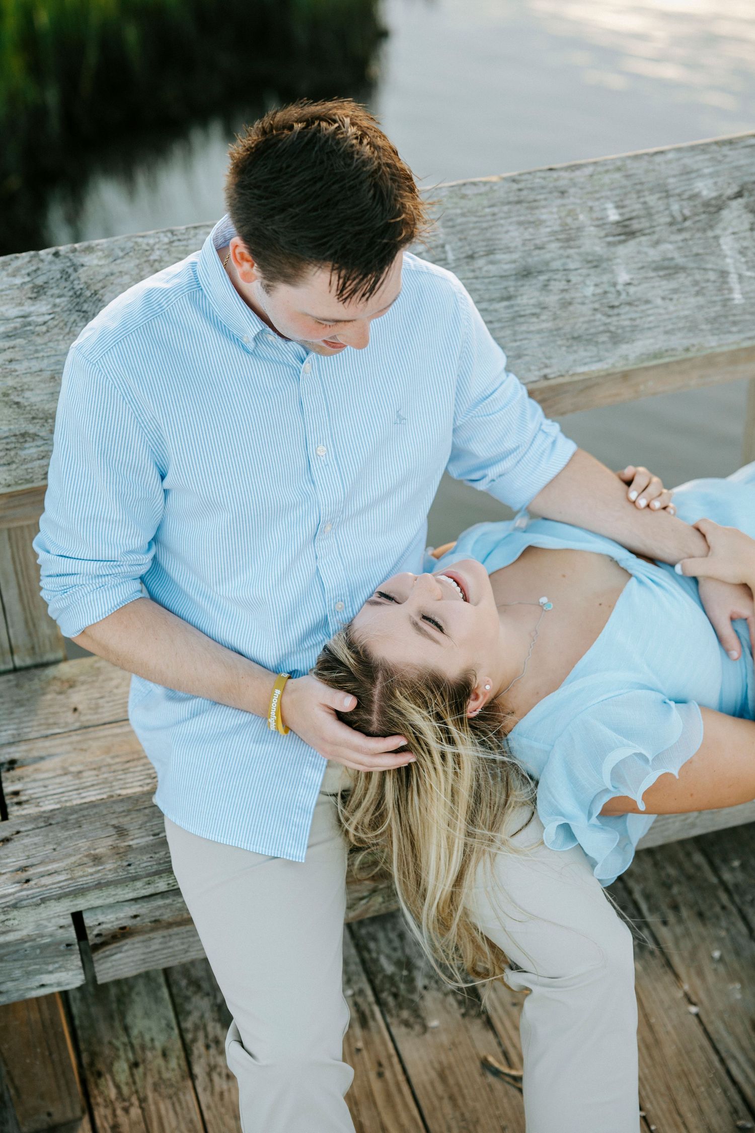 Romantic couple in light blue and khaki embracing on wooden dock by water at sunset.