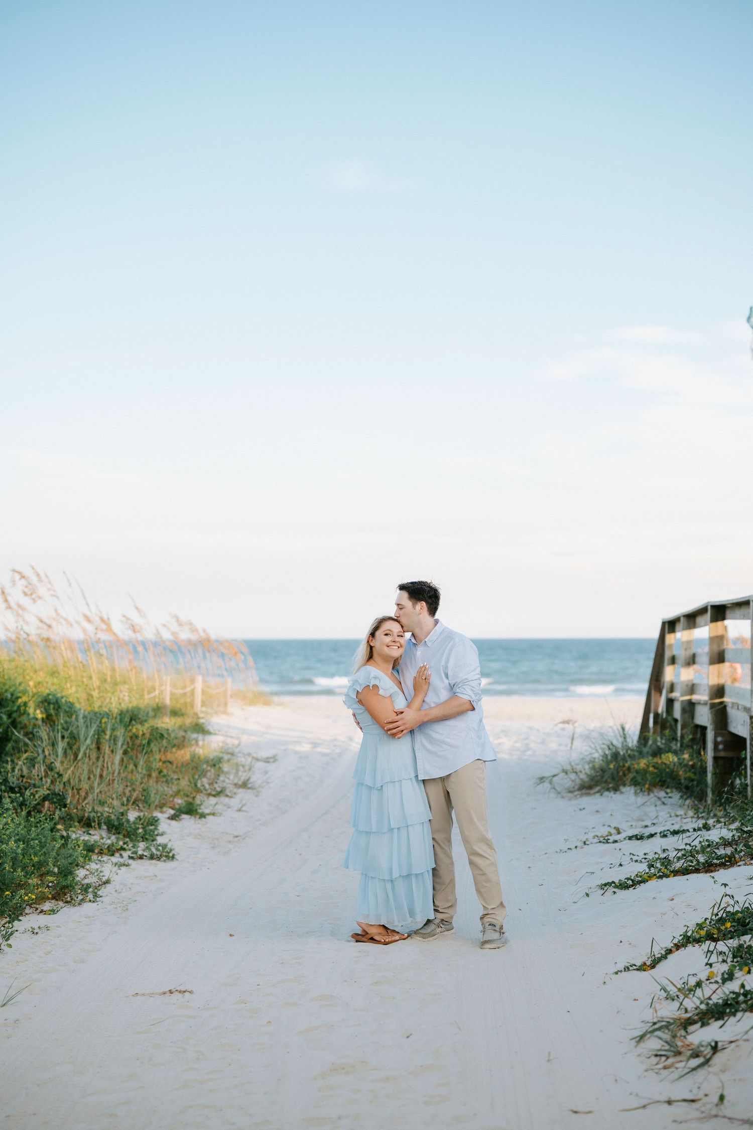 A romantic couple shares a tender moment on a sandy beach path with sea oats and ocean views at sunset.