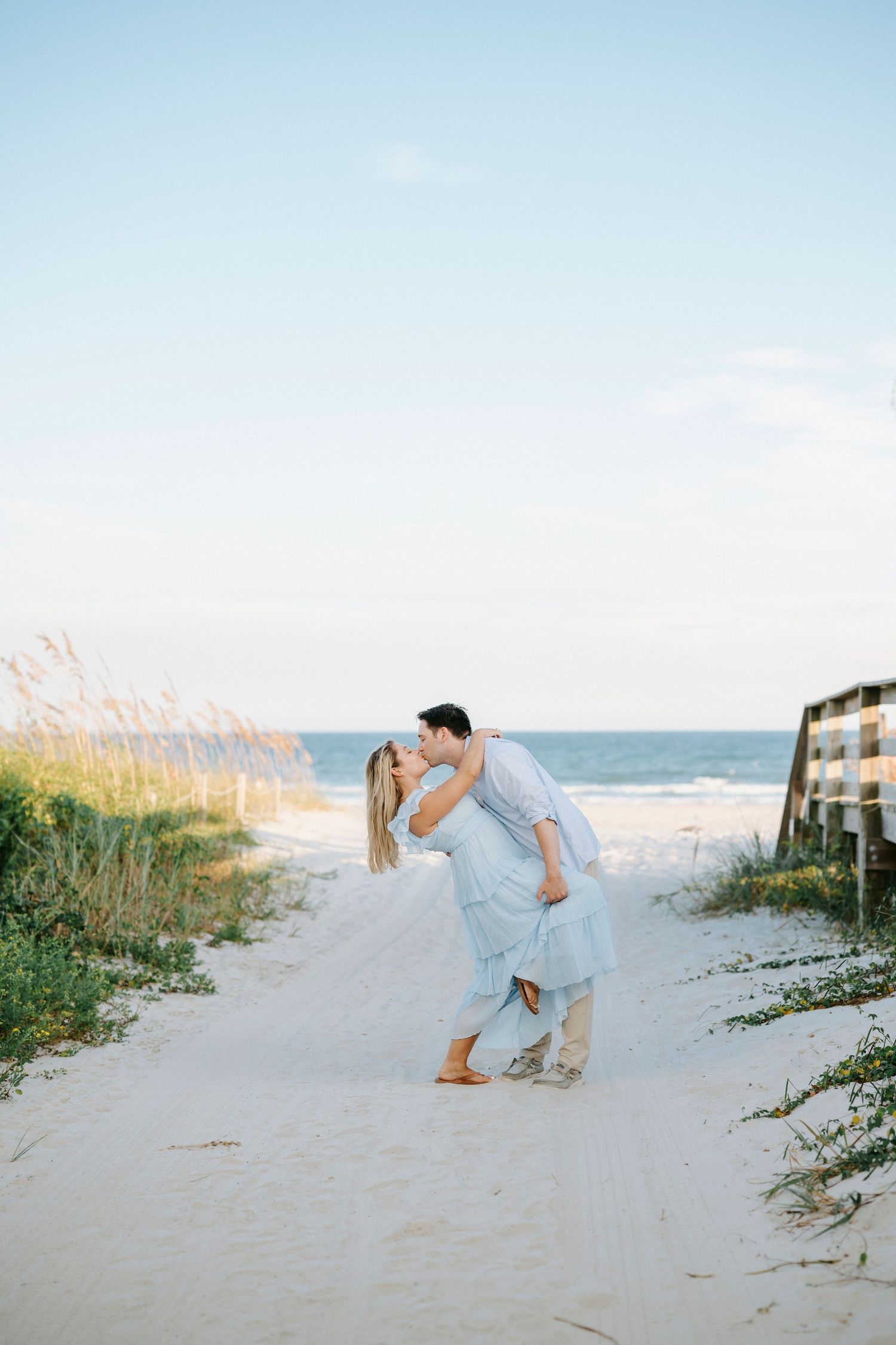 Couple in light blue outfits shares romantic moments on a sandy beach path with sea oats at sunset.