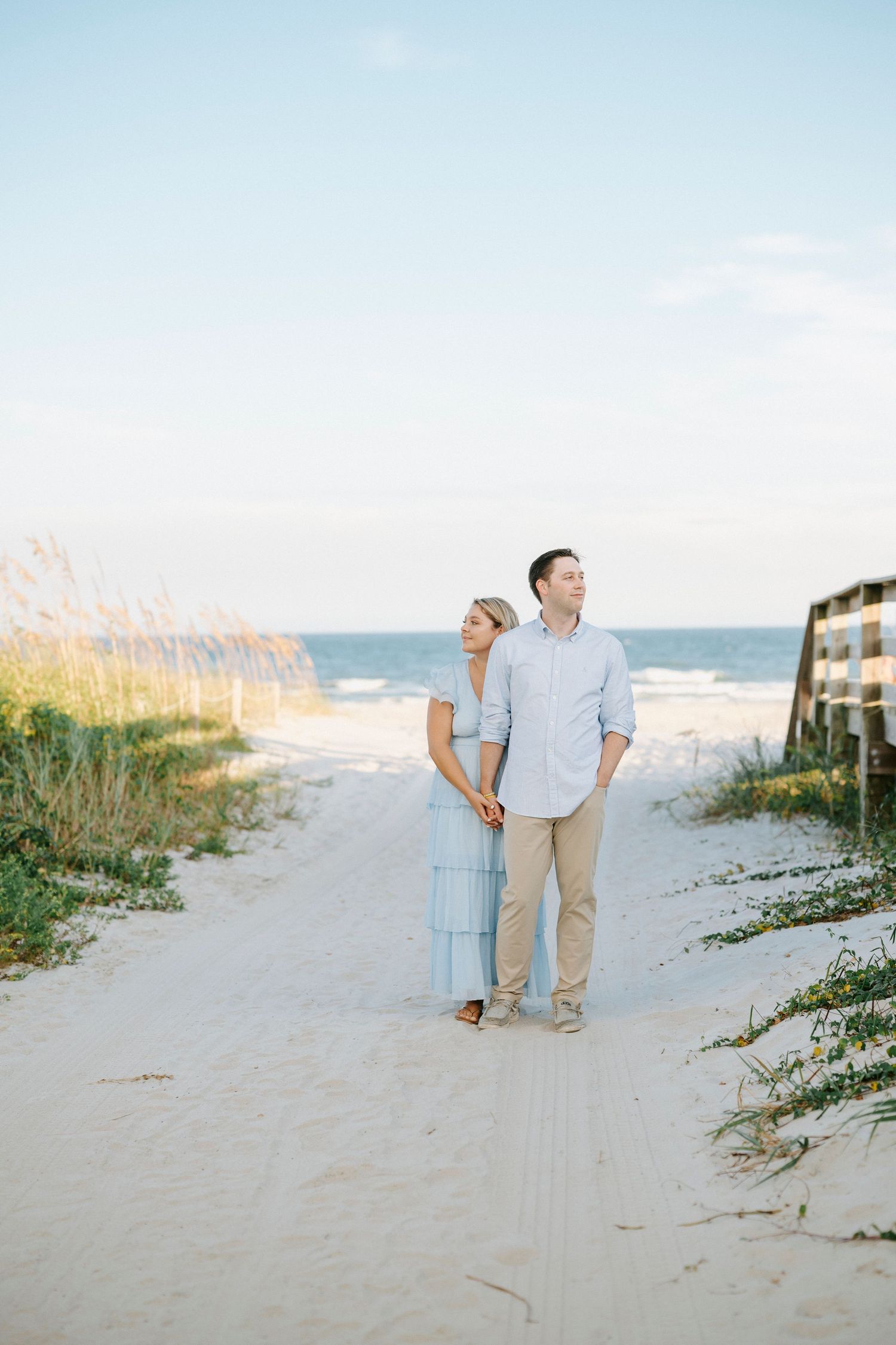 A couple in light blue and khaki attire walking together on a sandy beach path with ocean waves in the background.