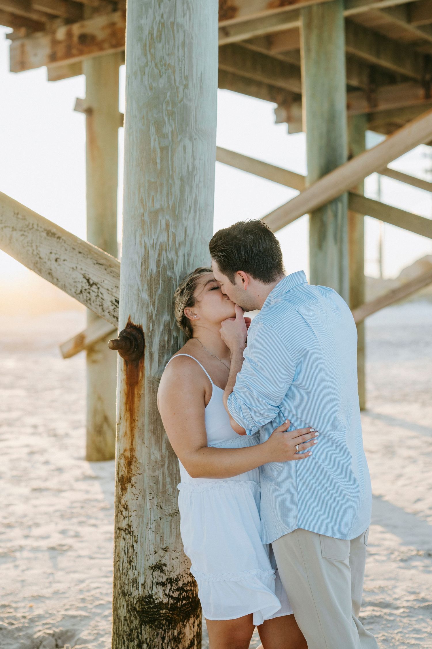A couple shares a tender moment under a wooden pier on a sandy beach during sunset.