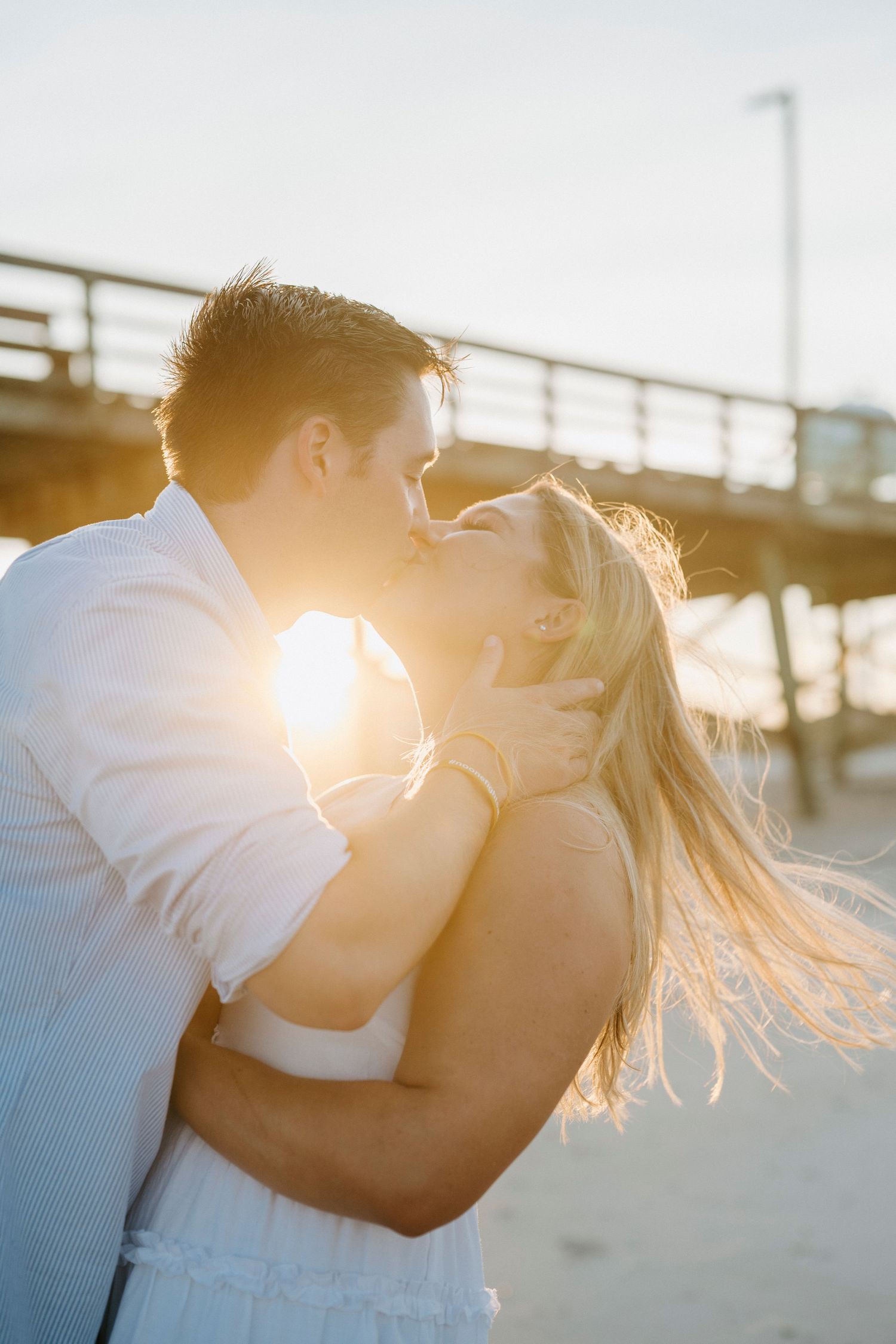 Romantic couple embracing and kissing on a pier during golden sunset hour with sunflare in background.