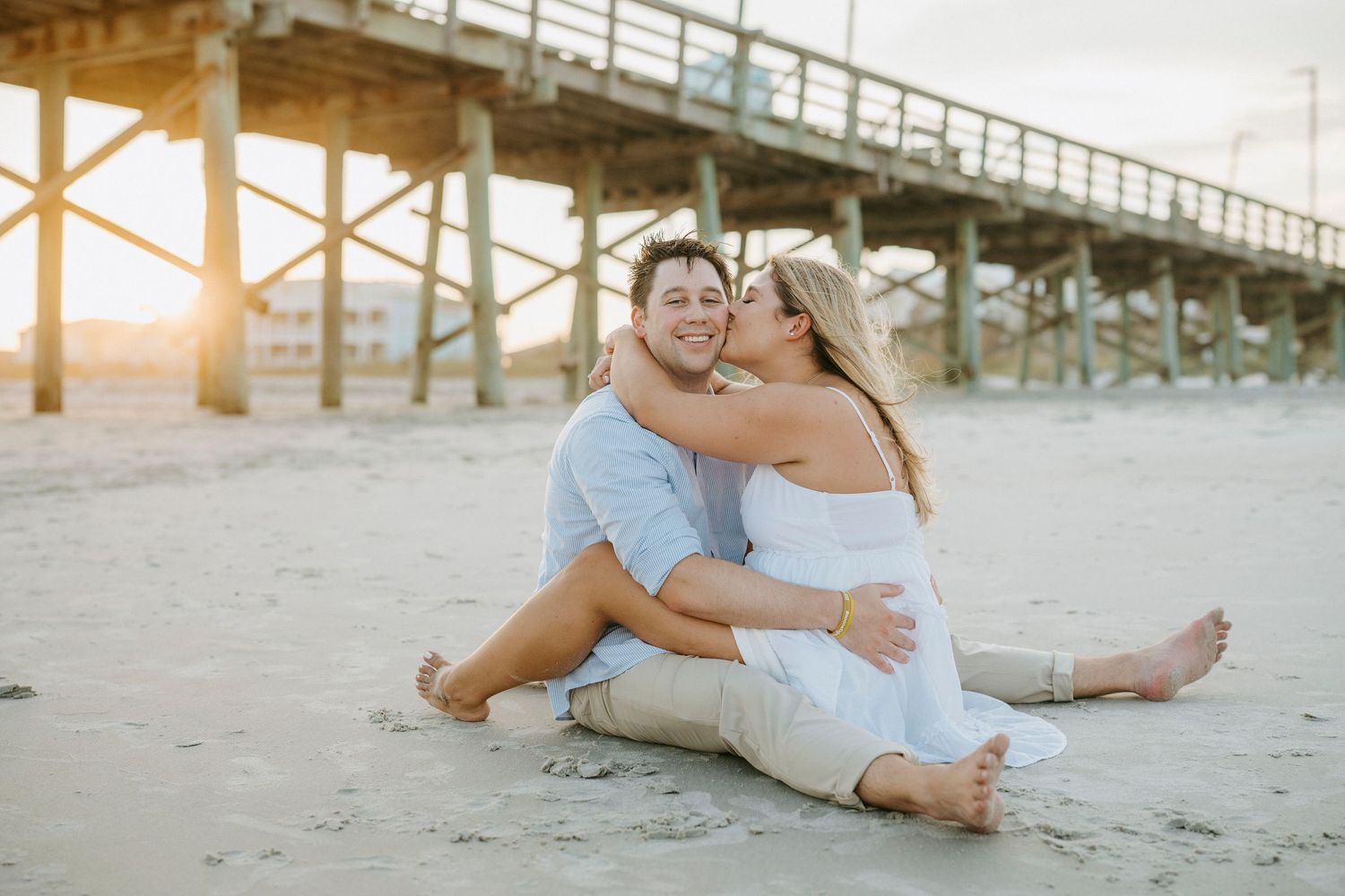 Couple embraces on sandy beach at sunset with wooden pier in background.