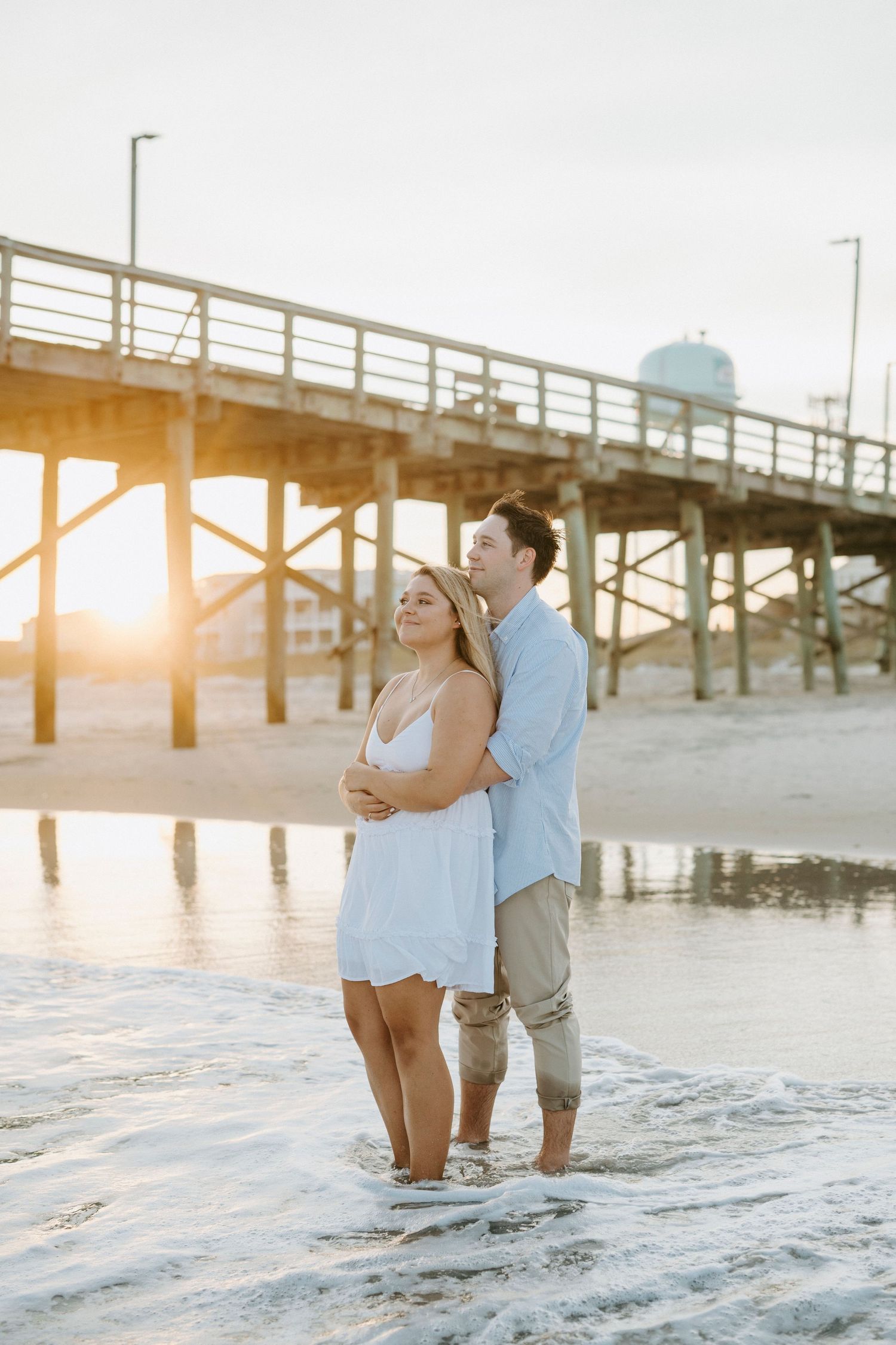 A couple stands together in the ocean waves at sunset with a wooden pier in the background.