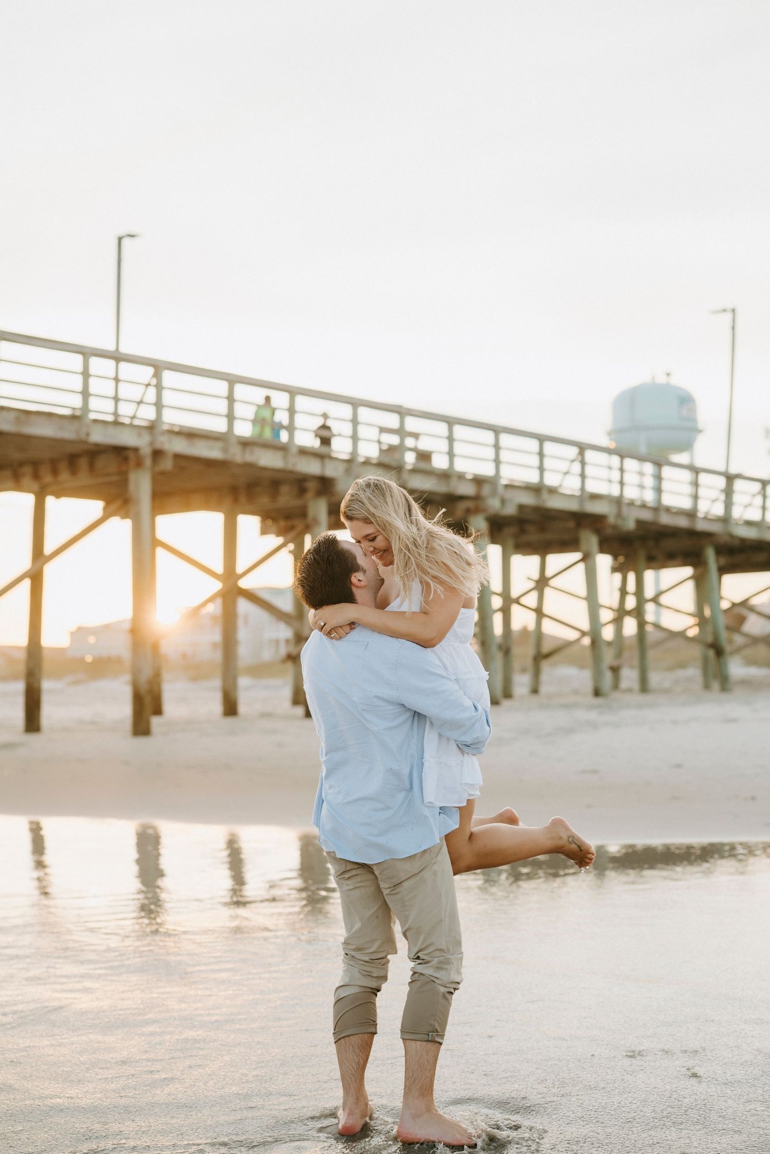 A couple embraces on a sandy beach at sunset near a wooden pier.