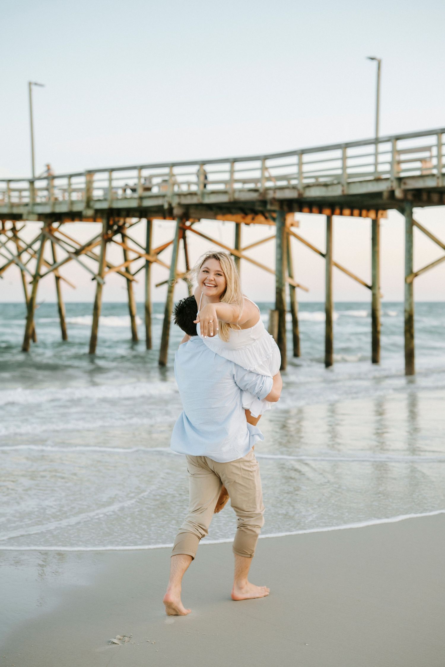 Playful couple dancing together barefoot on the beach with a wooden pier in the background.