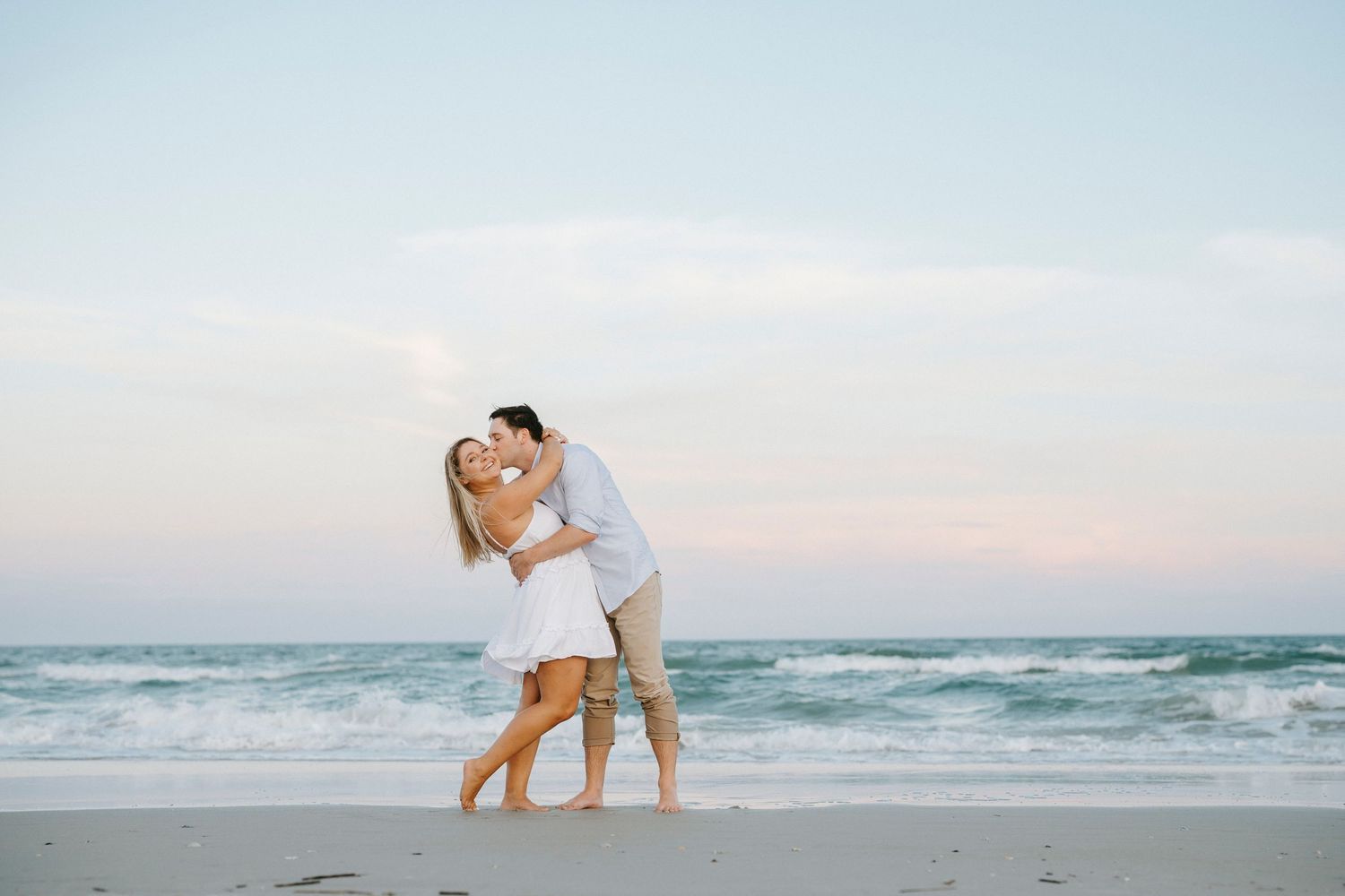 A couple shares a romantic moment on a beach at sunset with gentle waves rolling onto the shore.