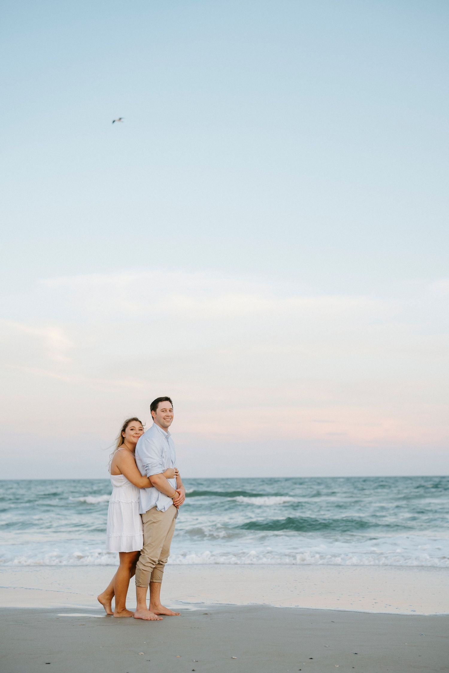 A couple shares a romantic moment together on a sandy beach at sunset with ocean waves in the background.