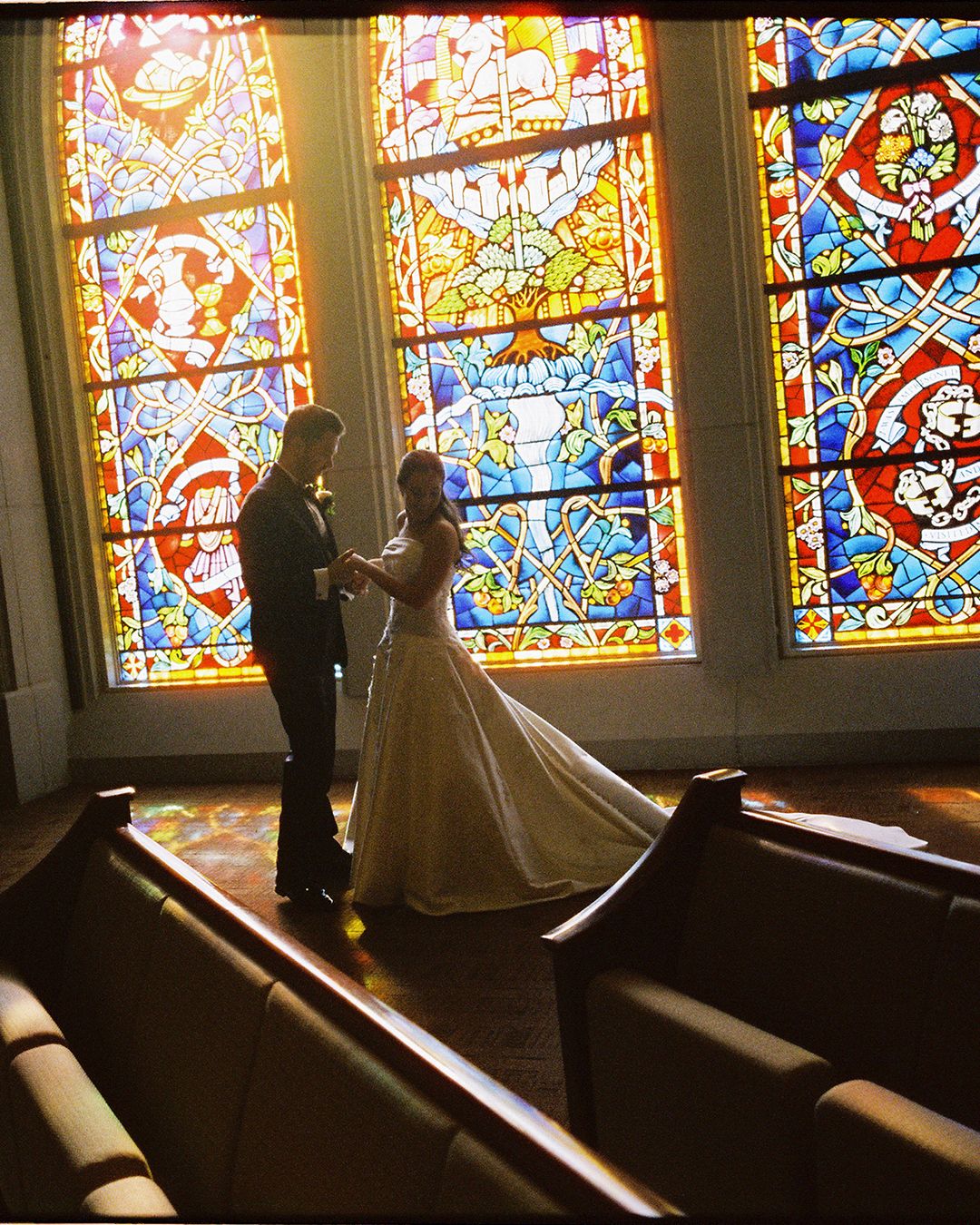 Silhouettes of a couple stand against vibrant stained glass windows in a church creating a dramatic backlit effect.
