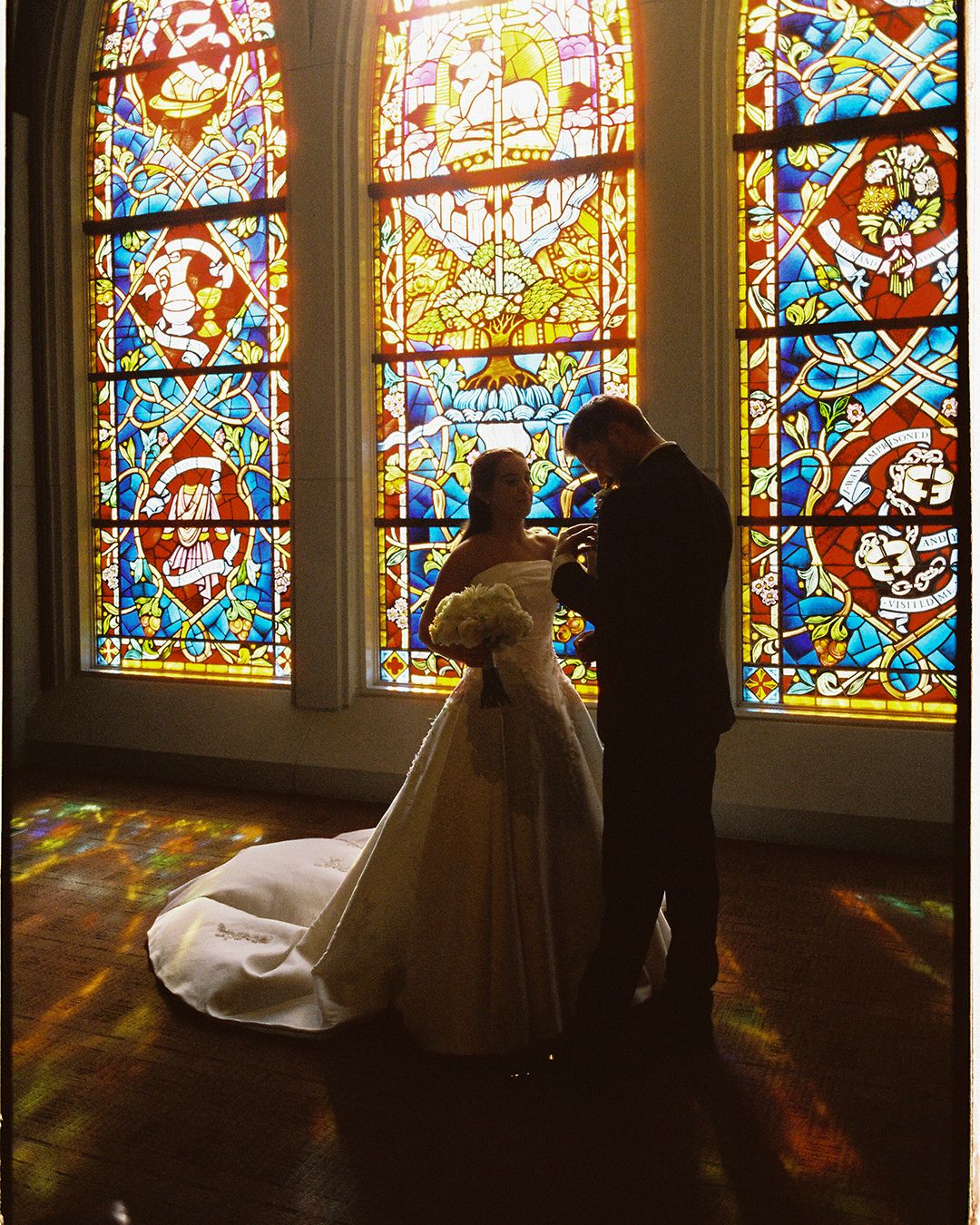 Silhouetted couple stands before vibrant stained glass church windows casting colorful reflections on the floor.