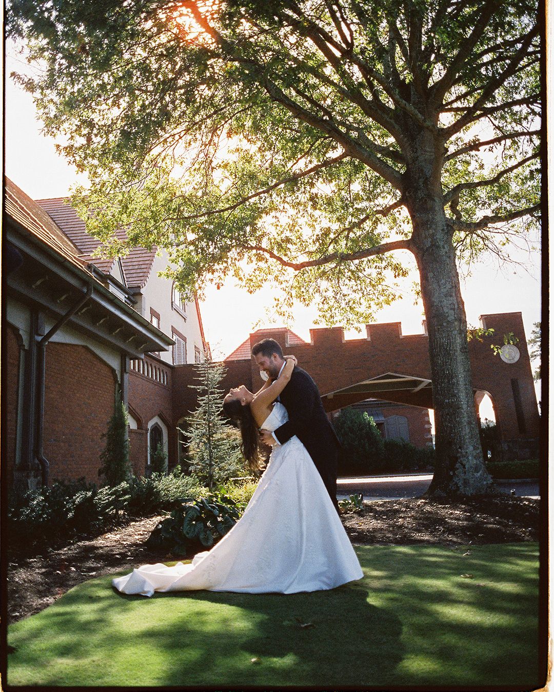 White wedding dress flows in motion against green lawn and brick church exterior in sunlight.