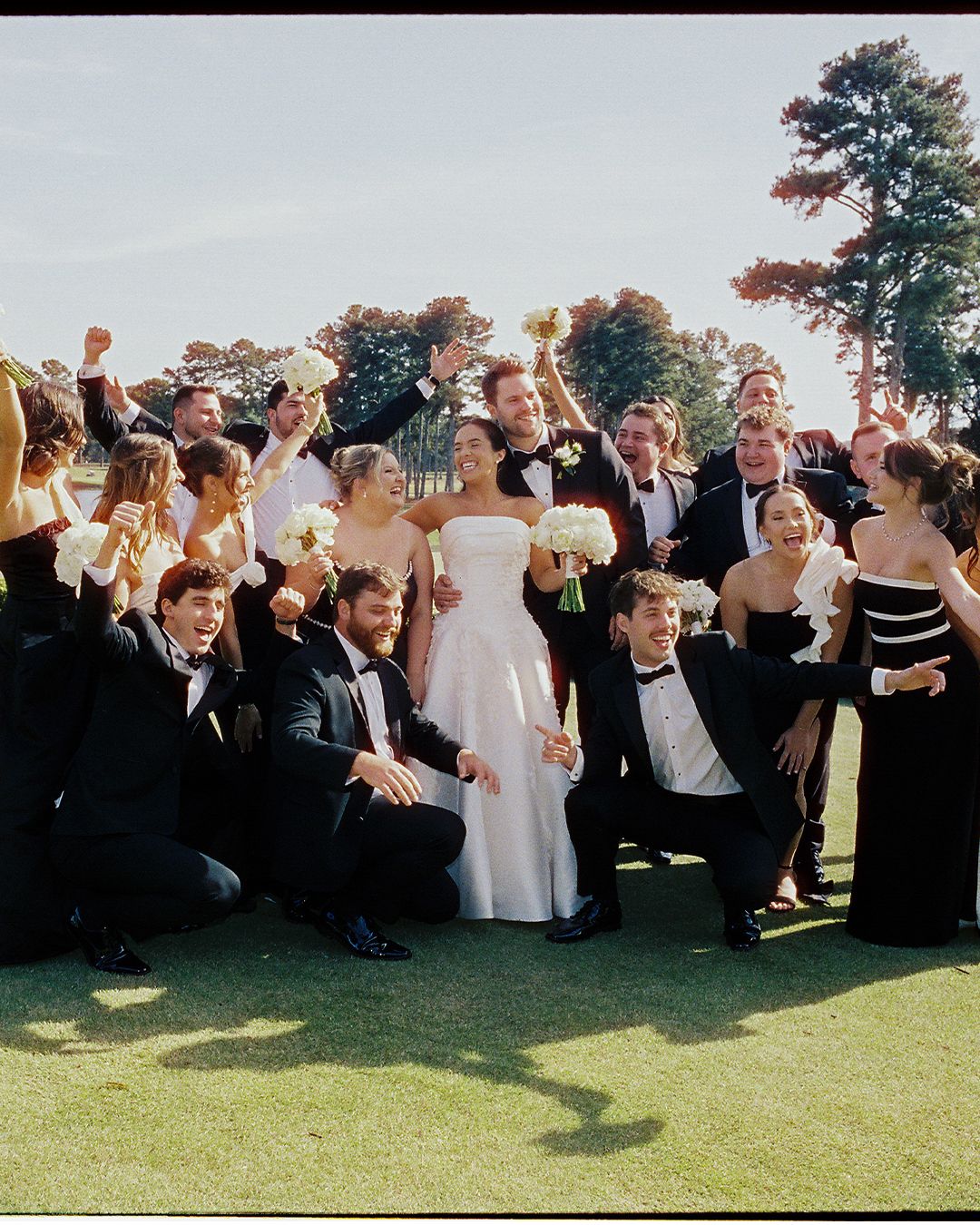 Wedding guests gathered on a lush green lawn cheer and celebrate during an outdoor reception.