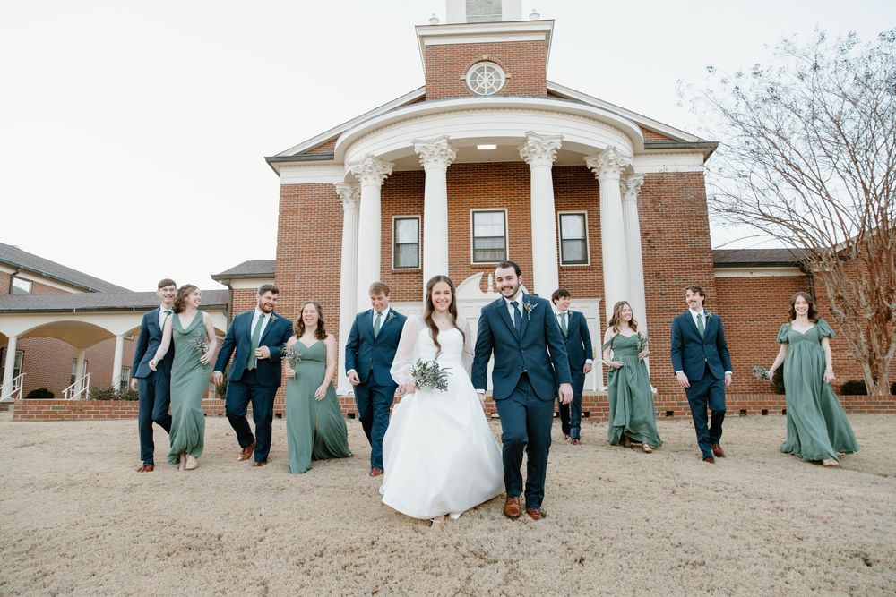 Wedding party in blue dresses and suits poses outside traditional brick church.