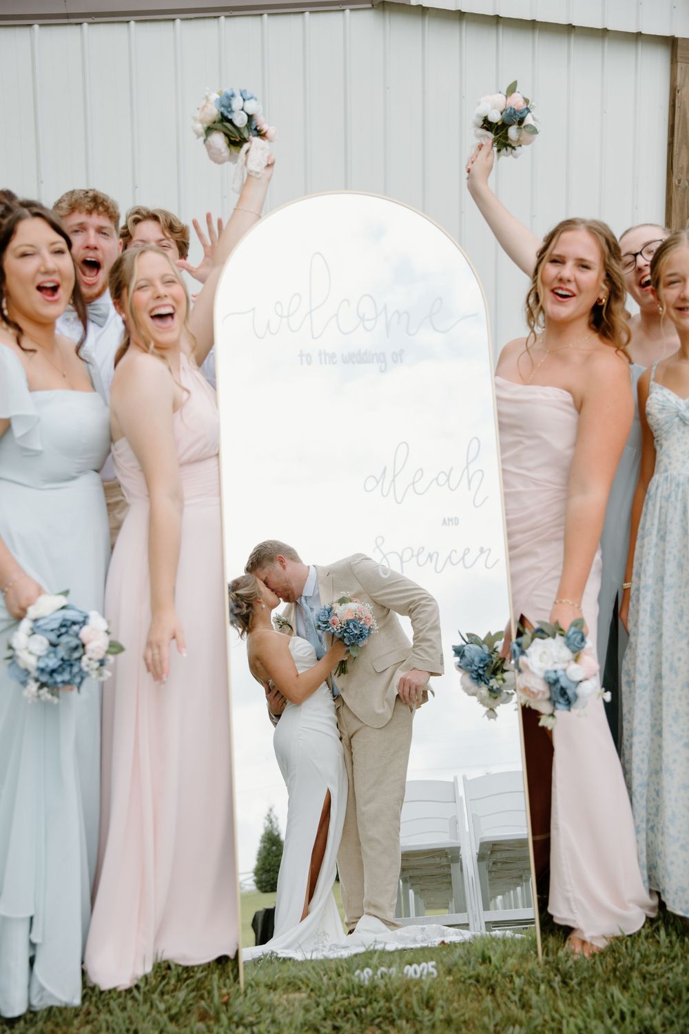 Wedding party members in pastel dresses and suits pose together against a white barn backdrop.