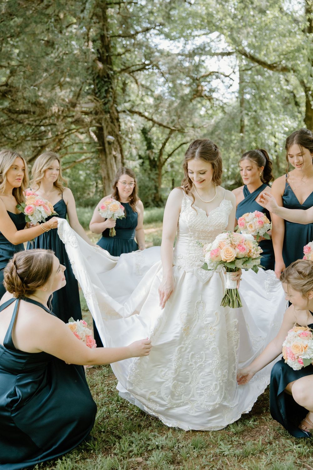 Bridesmaids in navy dresses help arrange the bride's white lace gown in an outdoor garden setting.