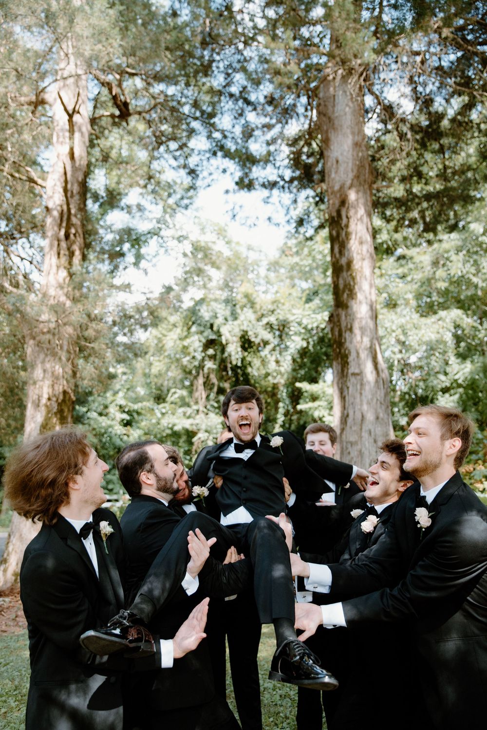 Groomsmen in black suits share drinks and laughter during outdoor wedding festivities.