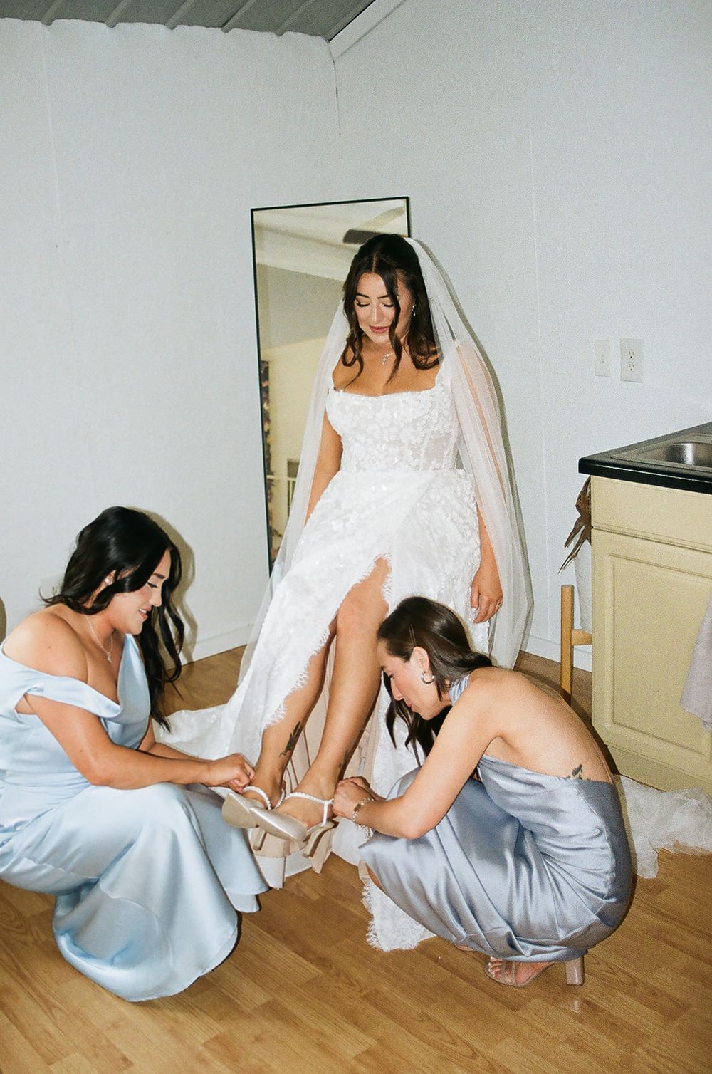 Two bridesmaids in blue satin dresses help the bride with her shoes before the wedding.