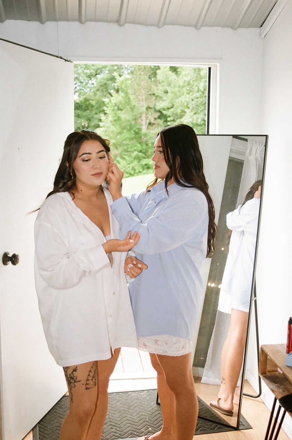 Two women in white shirts help each other with makeup preparations in a bright room.