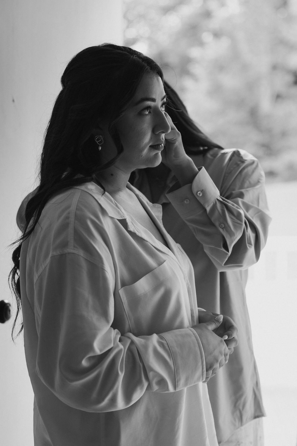 Black and white photo of a dark-haired bride in a white shirt touching her face before her wedding.