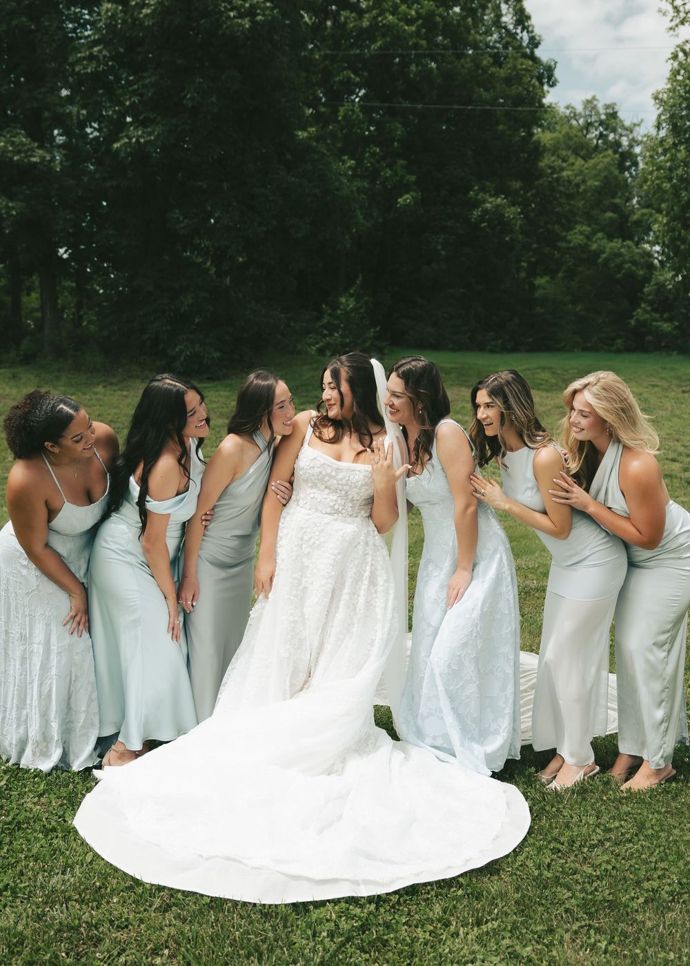 Bride in lace wedding gown laughs with six bridesmaids in mismatched sage green dresses on a green lawn.