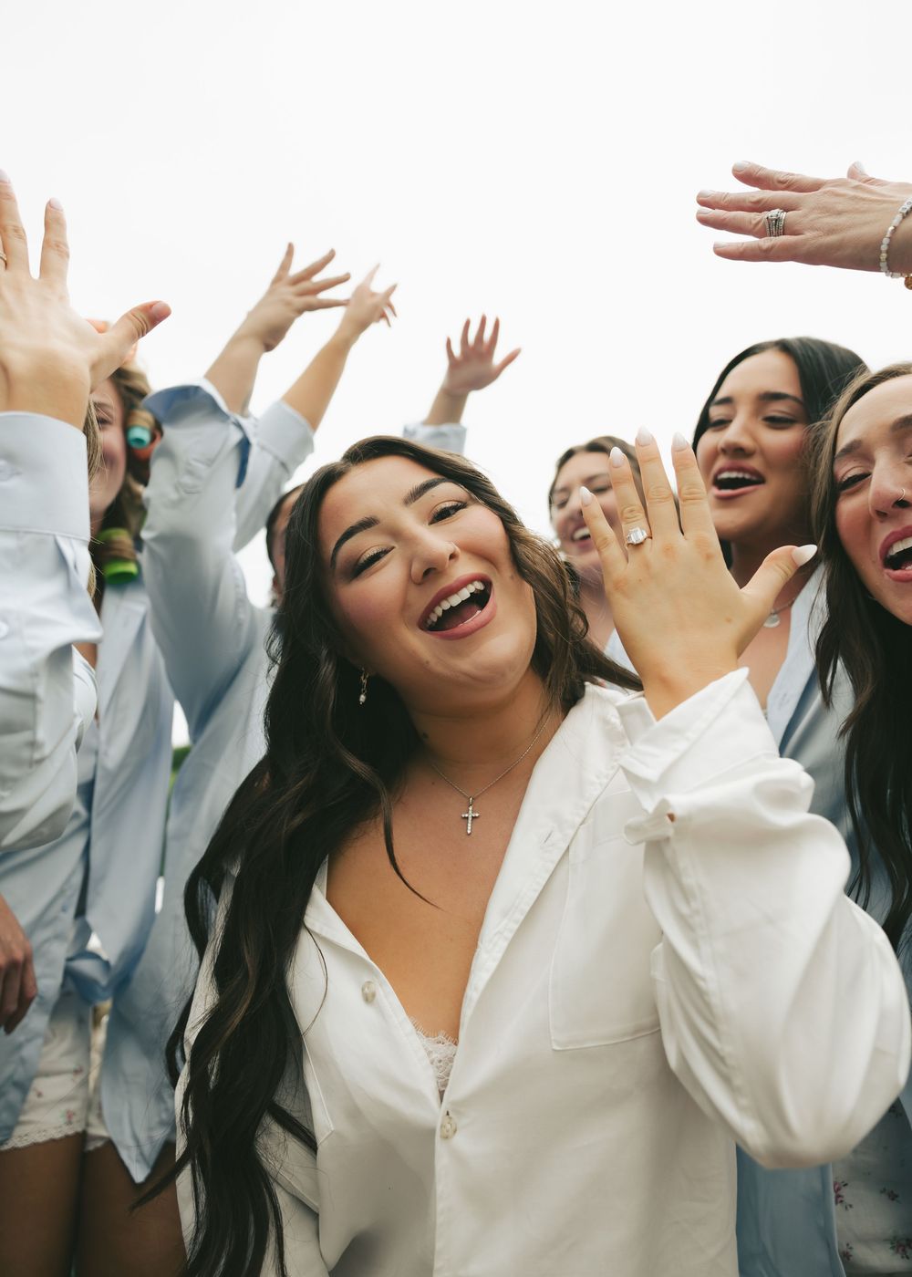 Joyful bride in white shirt showing engagement ring surrounded by cheering bridesmaids outdoors.