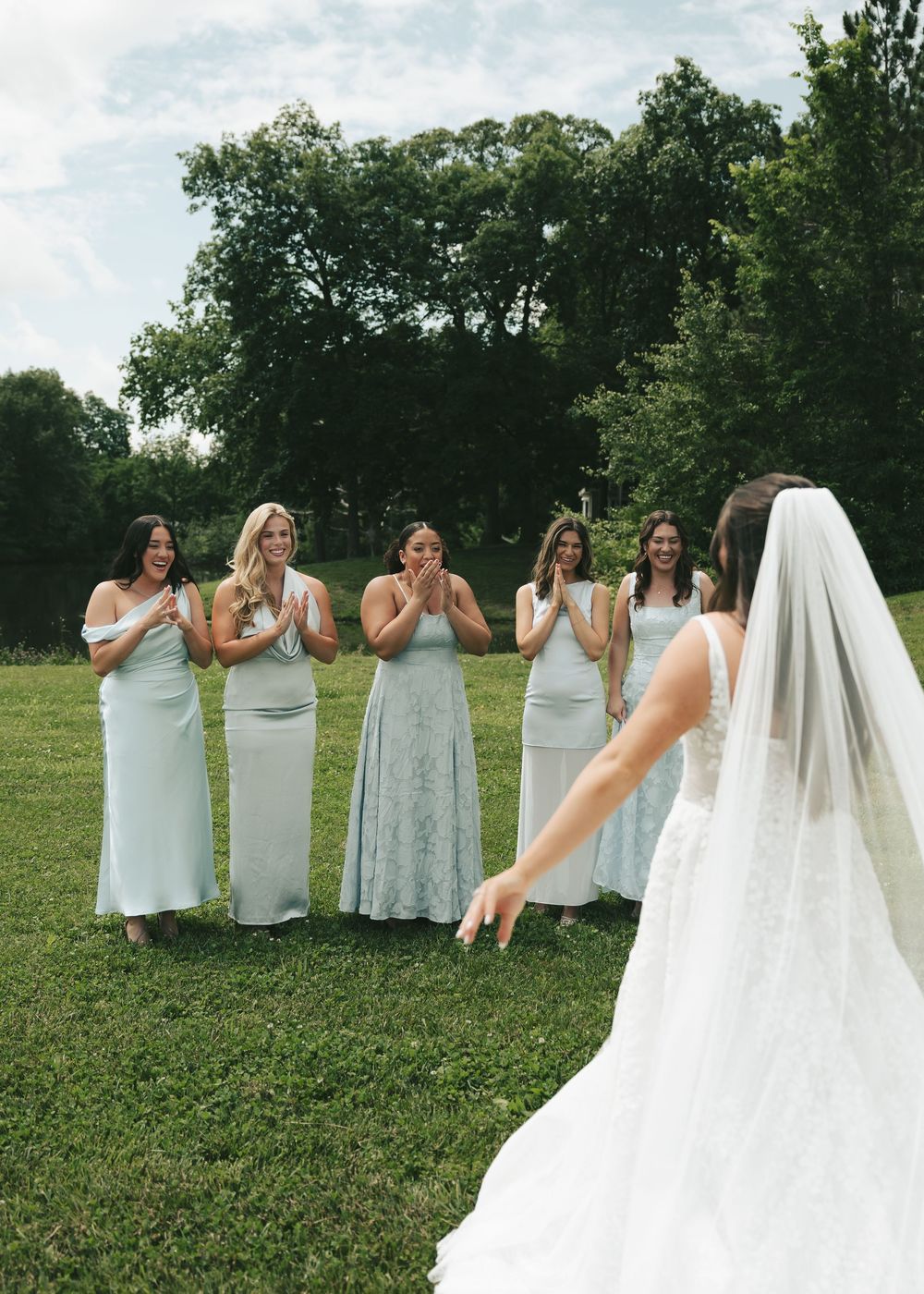 Bride in white lace gown surprises emotional bridesmaids in sage green dresses on green lawn.