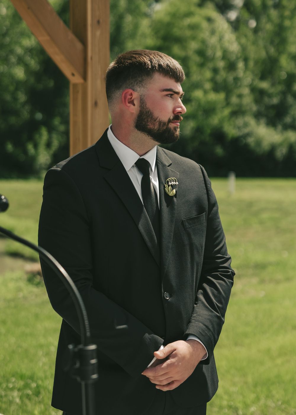 Handsome groom in black suit with boutonniere waits at the altar during outdoor ceremony.