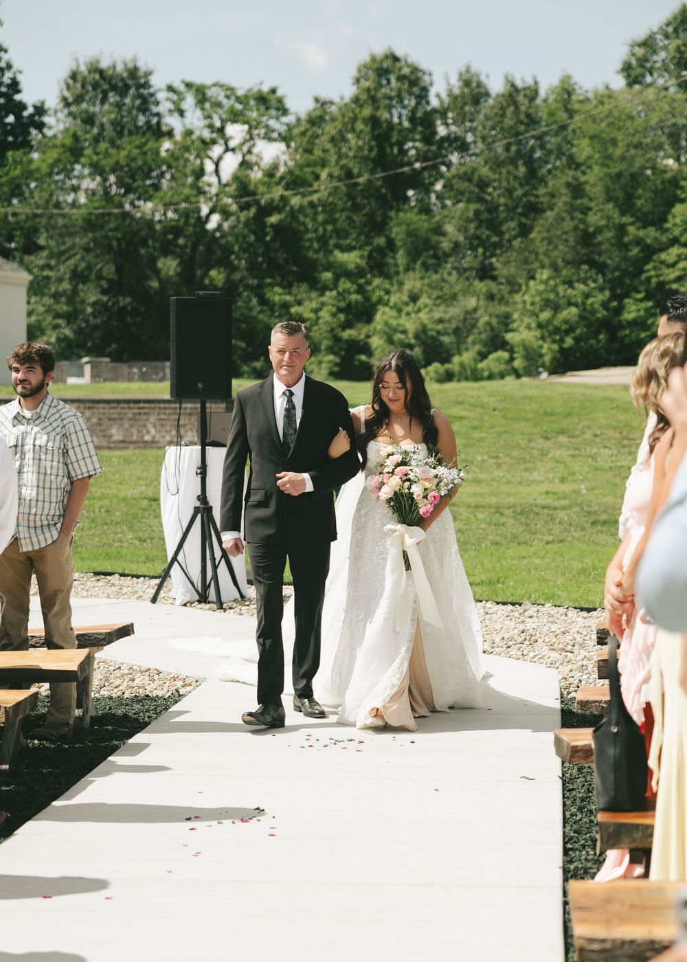 Father escorts bride holding pink bouquet down outdoor wedding aisle lined with guests.