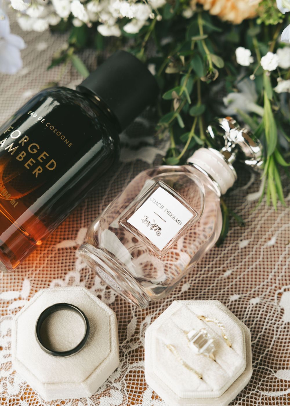 Flat lay of two perfume bottles, wedding rings in a velvet box, and white florals on a lace background.