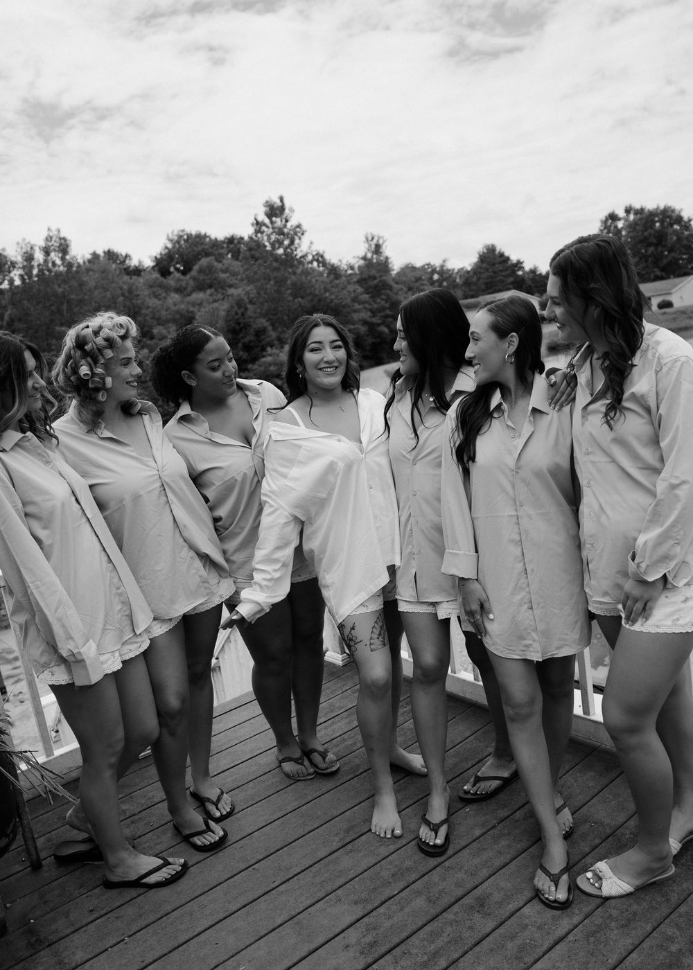 Black and white photo of bride and bridesmaids laughing together on a deck wearing matching white button-down shirts.