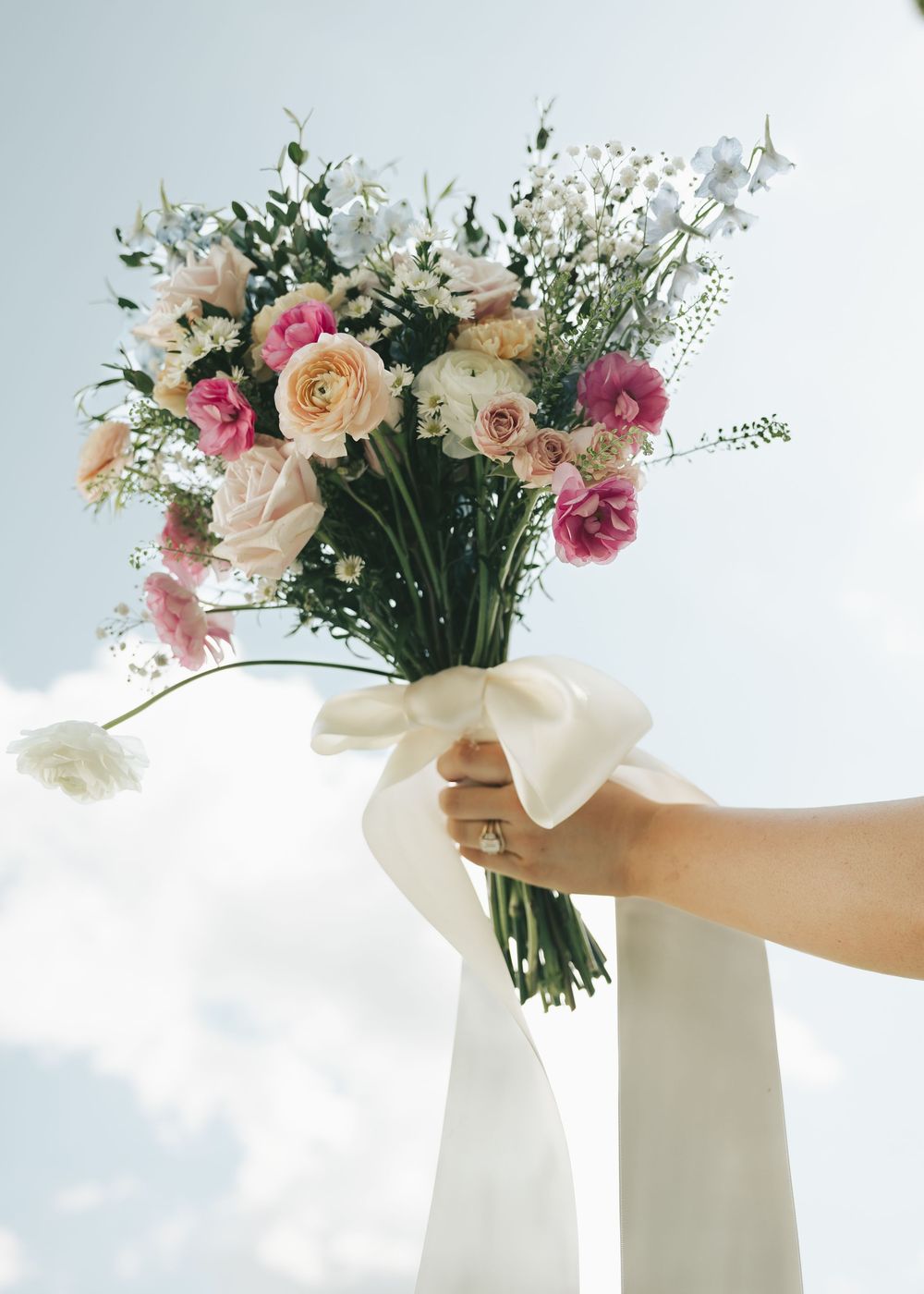 Hand holds elegant bridal bouquet of pink, white, and peach flowers with a long white ribbon.