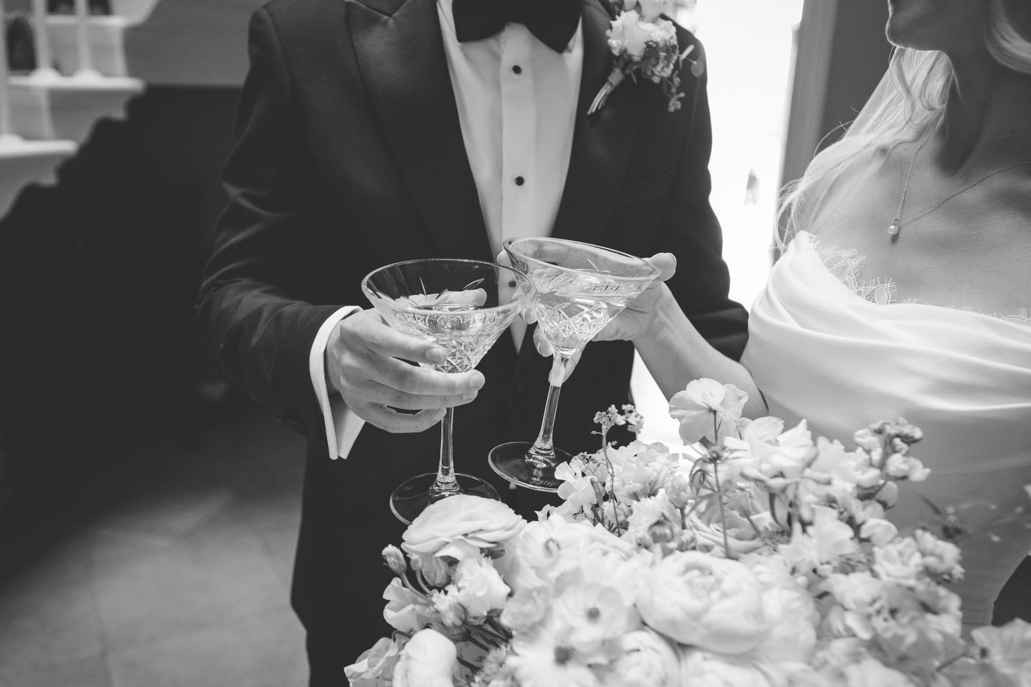A black and white photograph shows a bride in a flowing white gown with a train holding a bouquet in a shadowy doorway.