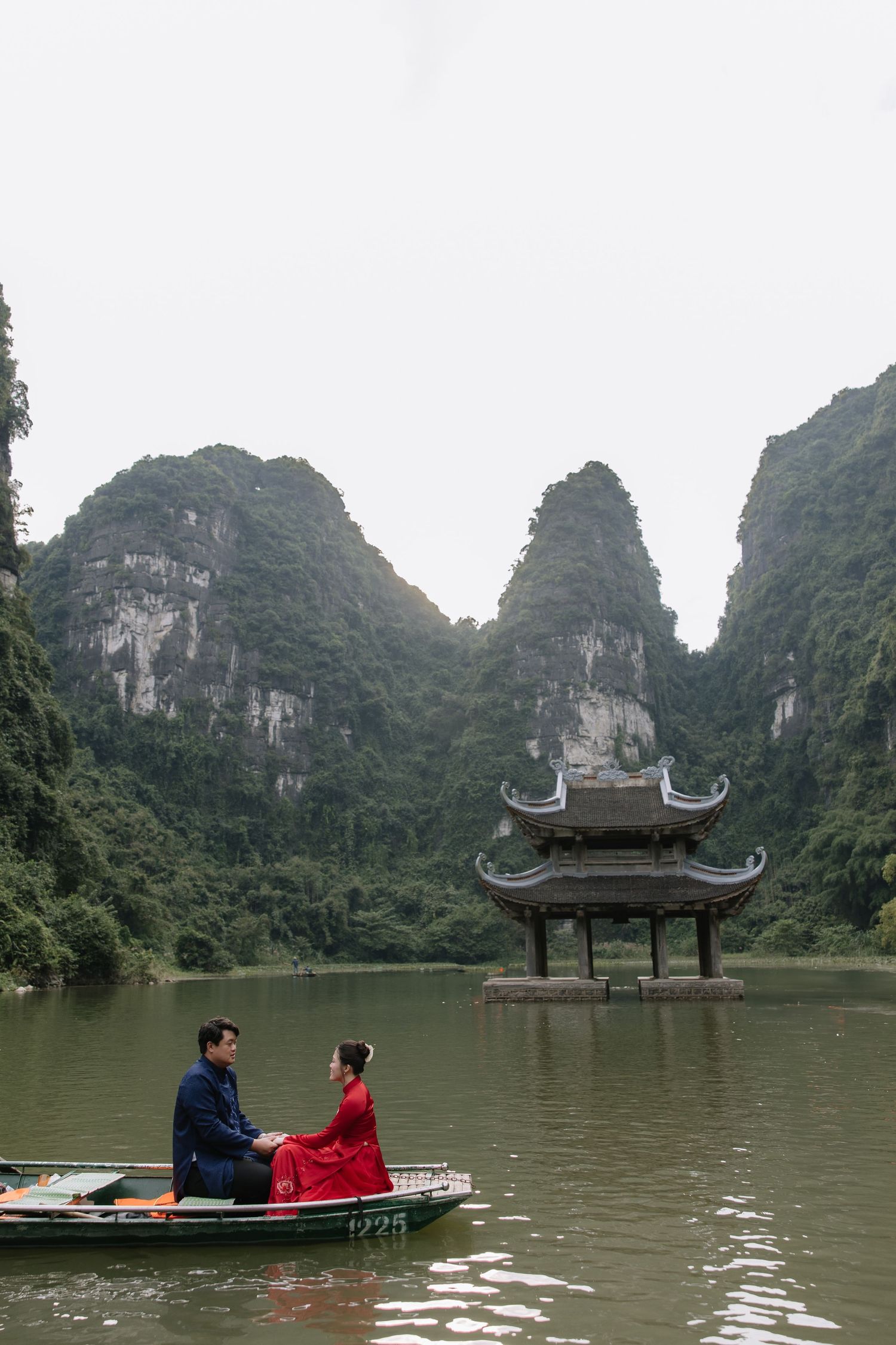 Couple in traditional Vietnamese attire sitting in a boat near a pagoda amid karst mountains.