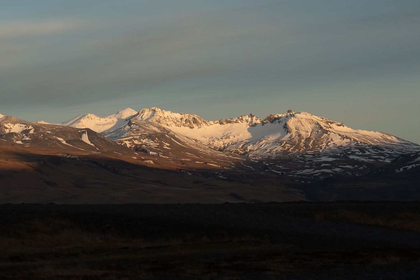 Snow-capped mountain range glowing in golden hour light under a dramatic cloudy sky.