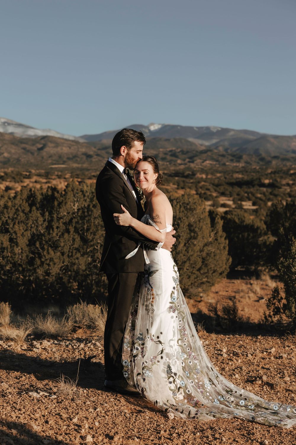 Couple embracing in wedding attire with snow-capped mountains and desert scenery behind them.