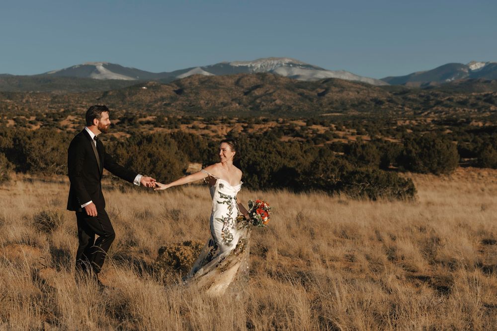 Bride and groom holding hands walking through golden meadow with mountain backdrop.