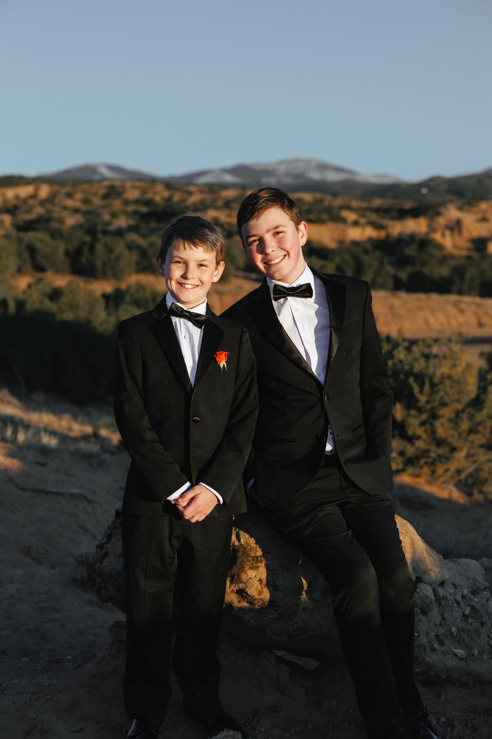 Two boys in black tuxedos with bow ties pose on rocks with a desert landscape at sunset.