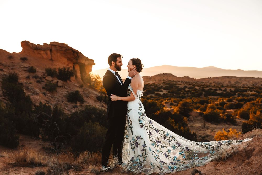 Bride in floral gown and groom embrace at sunset among red rock desert formations.