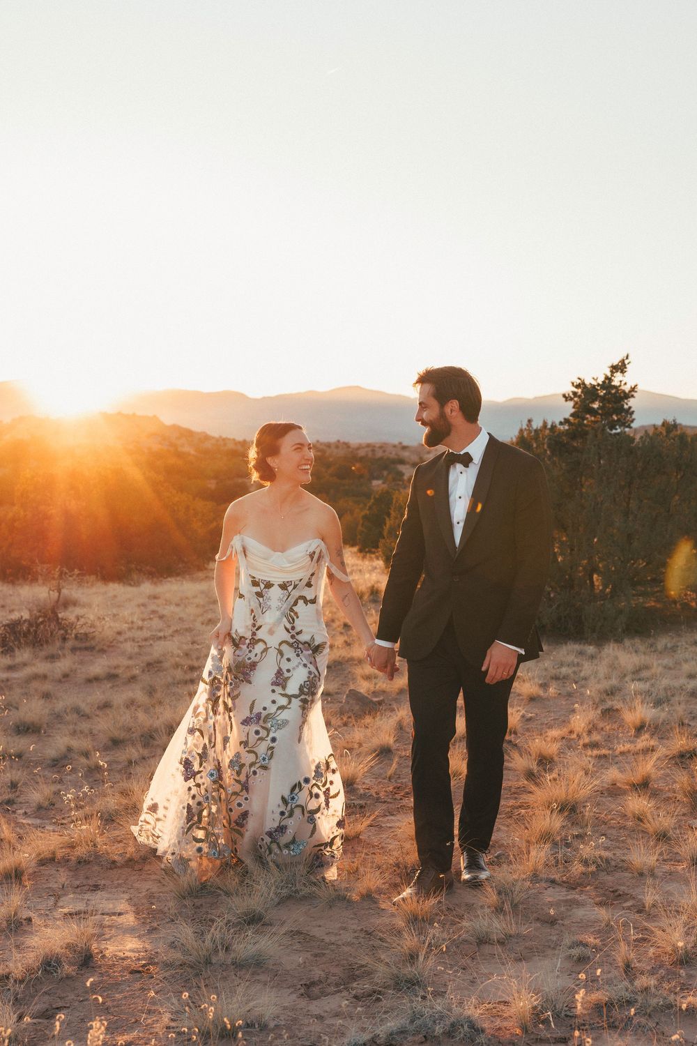 Bride and groom walk hand in hand through desert field during a stunning golden sunset.