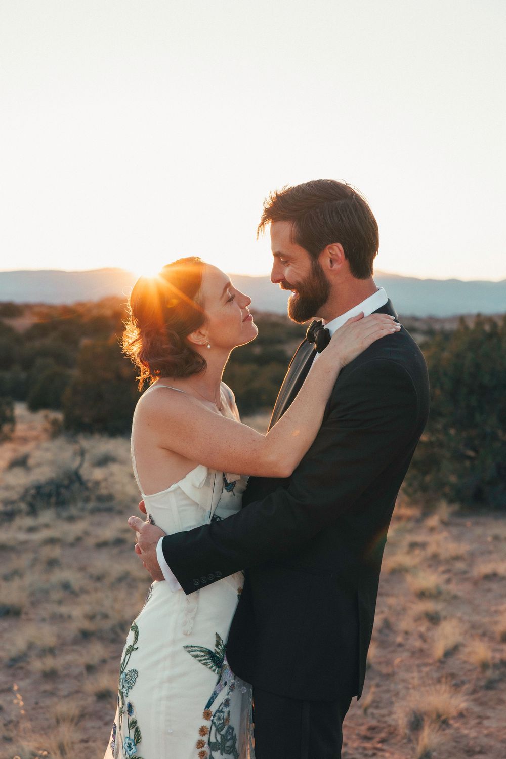 Bride and groom embrace tenderly at golden hour with a radiant desert sunset behind them.