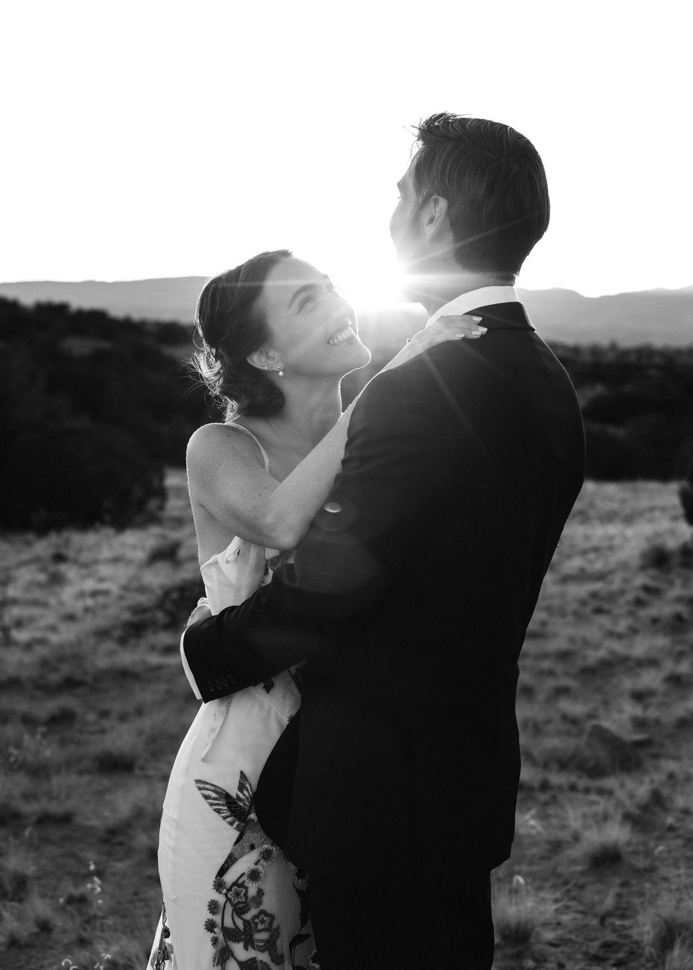Black and white photo of bride and groom embracing with sunburst between them in a field.